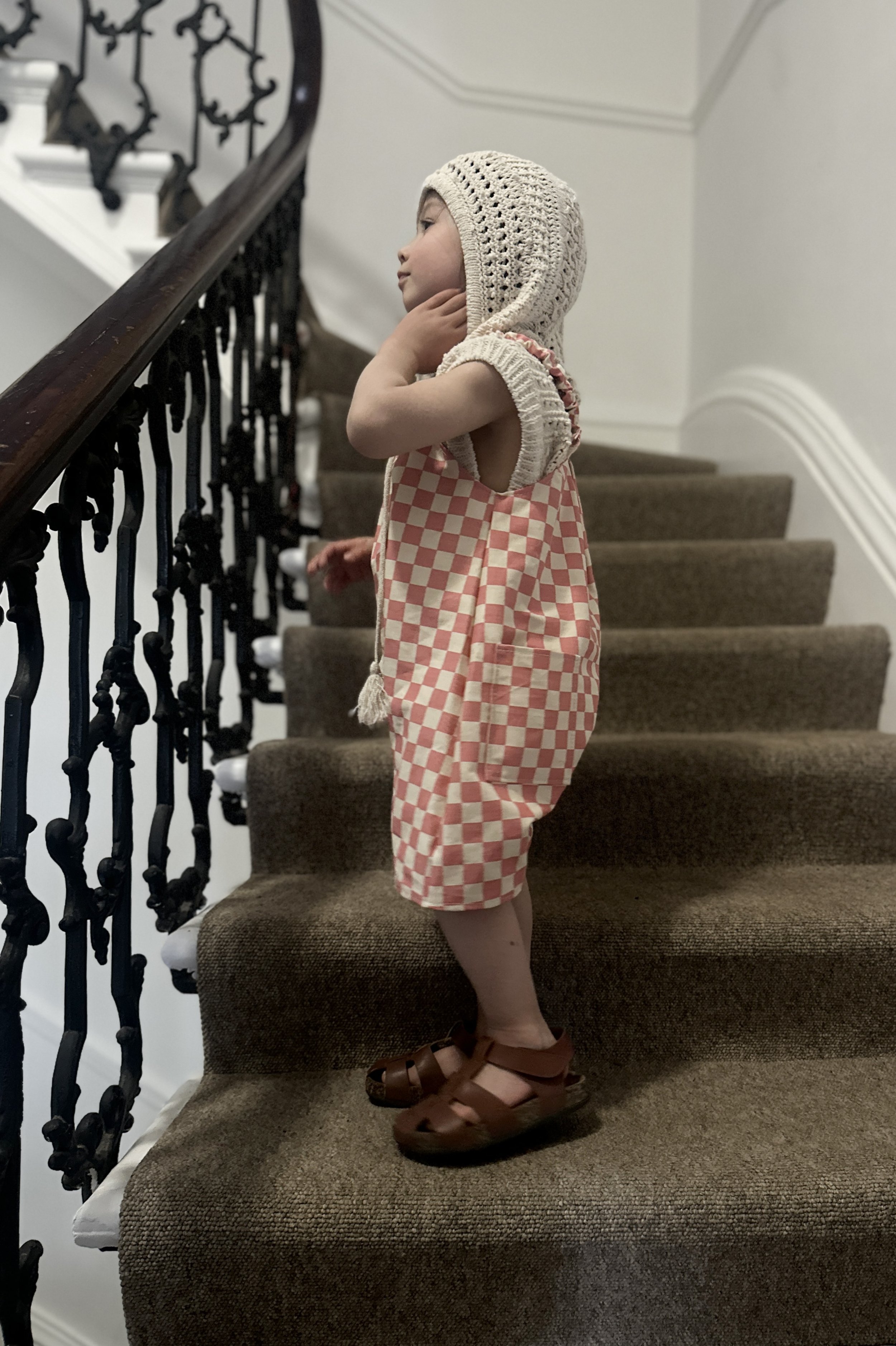 side view of child wearing organic cotton canvas pullover short dungarees in pink checkerboard colour, layered over crochet short sleeves hoodie top, brown sandals, standing on staircase