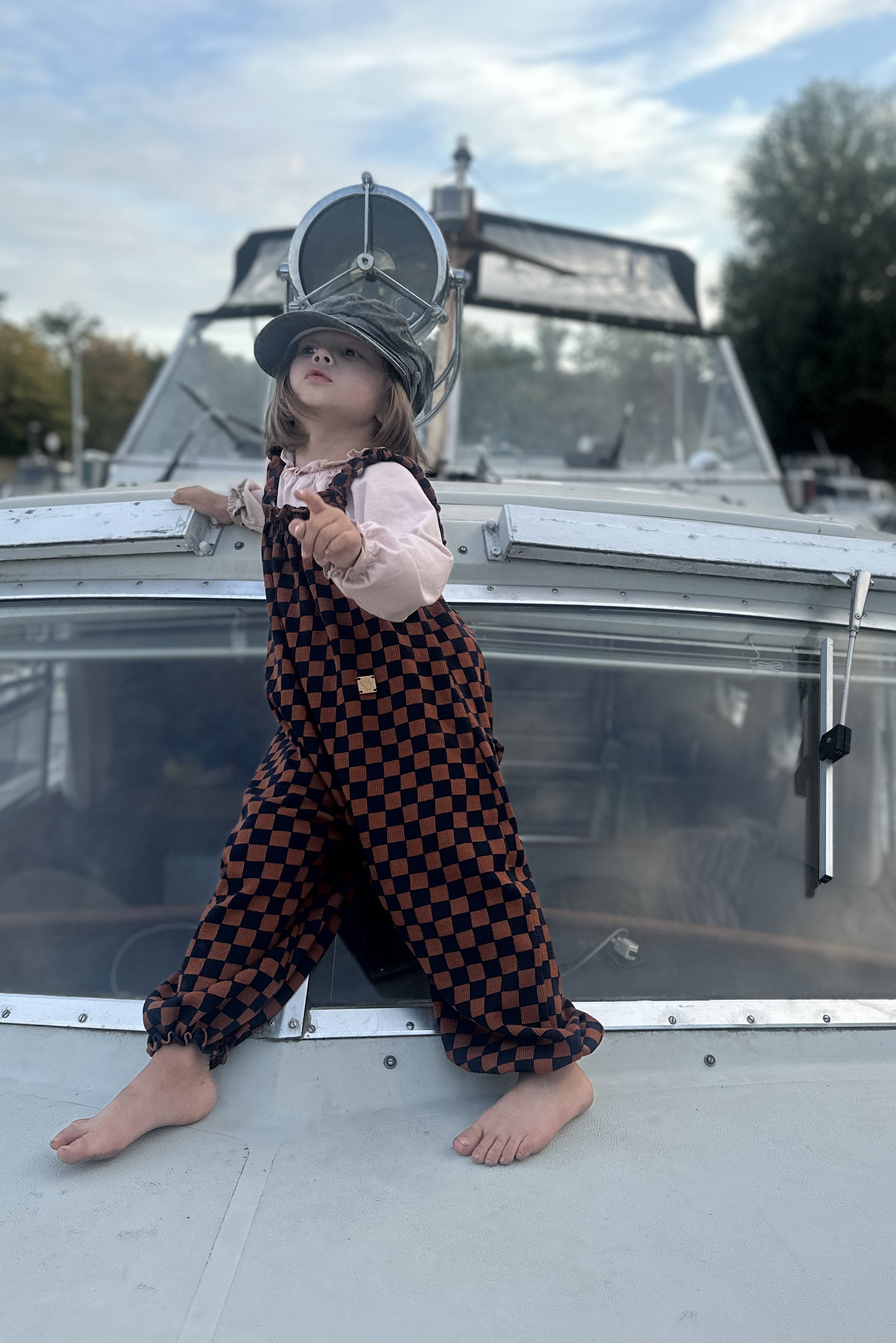 child wearing pullover dungarees in checkerboard navy and rust colour, standing on a boat at marina