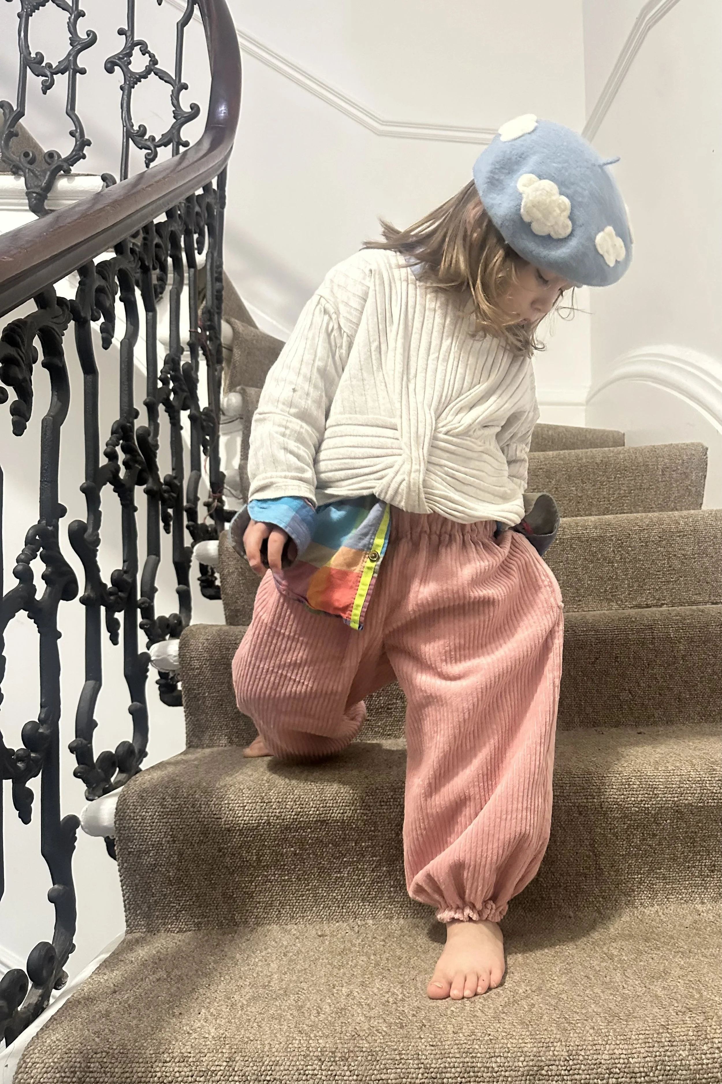 child wearing organic cotton corduroy pink trousers barrel shape baggy fit oversized elastic hem, with white top, rainbow shirt, blue cloudy hat, standing on staircase