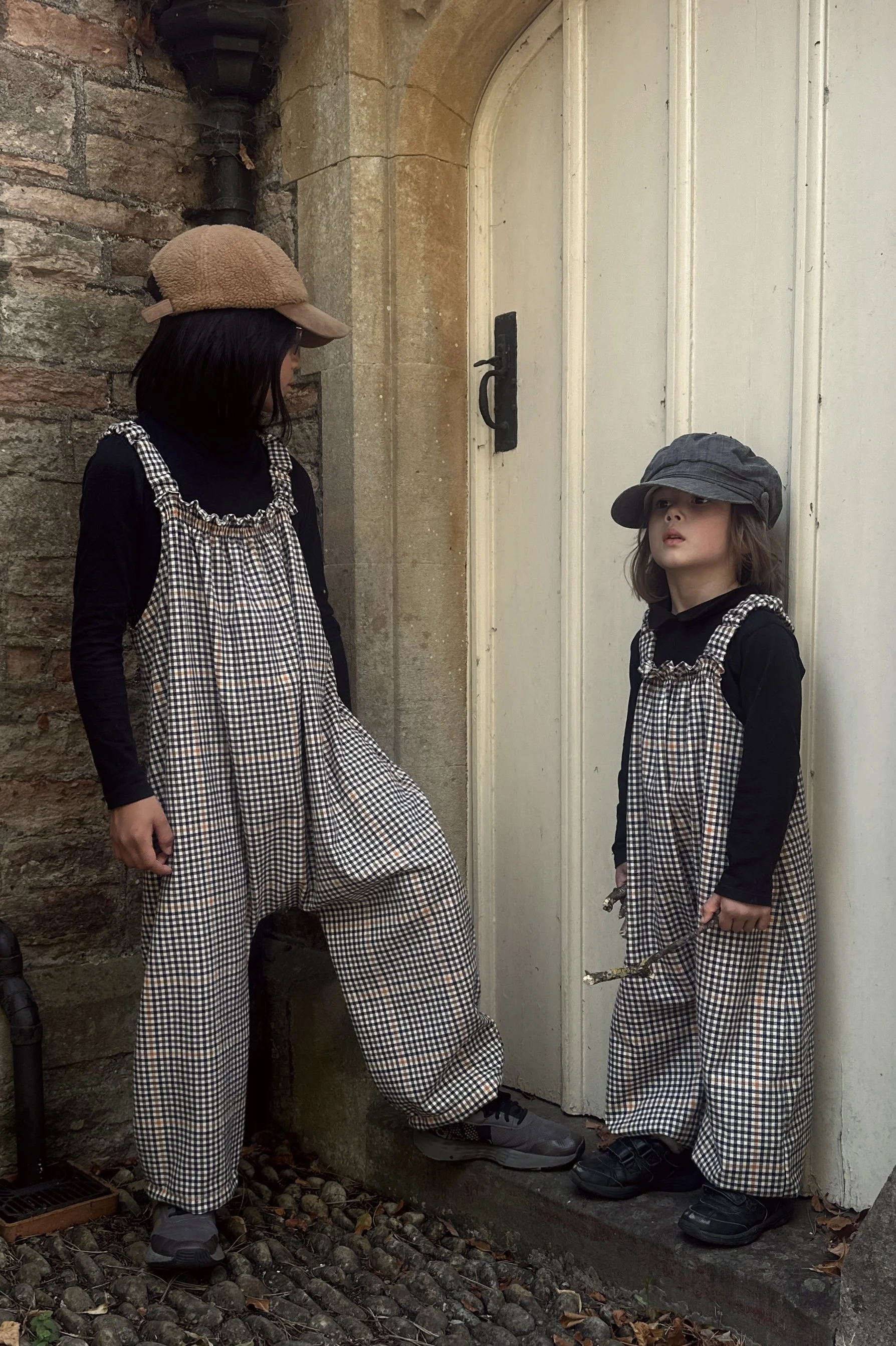 a child and an adult wearing organic cotton retro brown check dungarees layered with black top, grey hat, standing in front of a white doo
