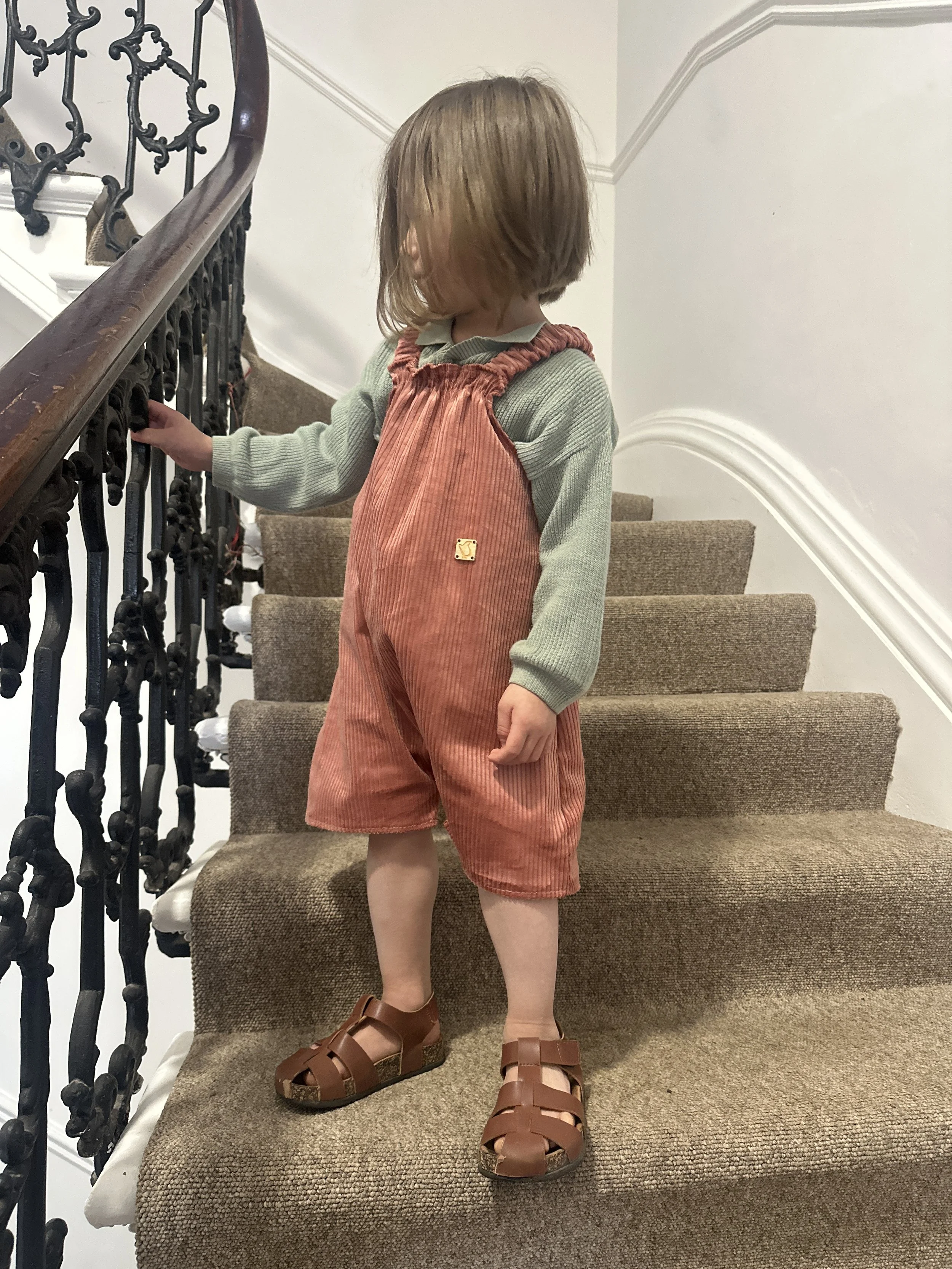child wearing organic cotton corduroy short dungarees coral colour, layered over a duck egg green jumper, brown sandals, standing on staircase