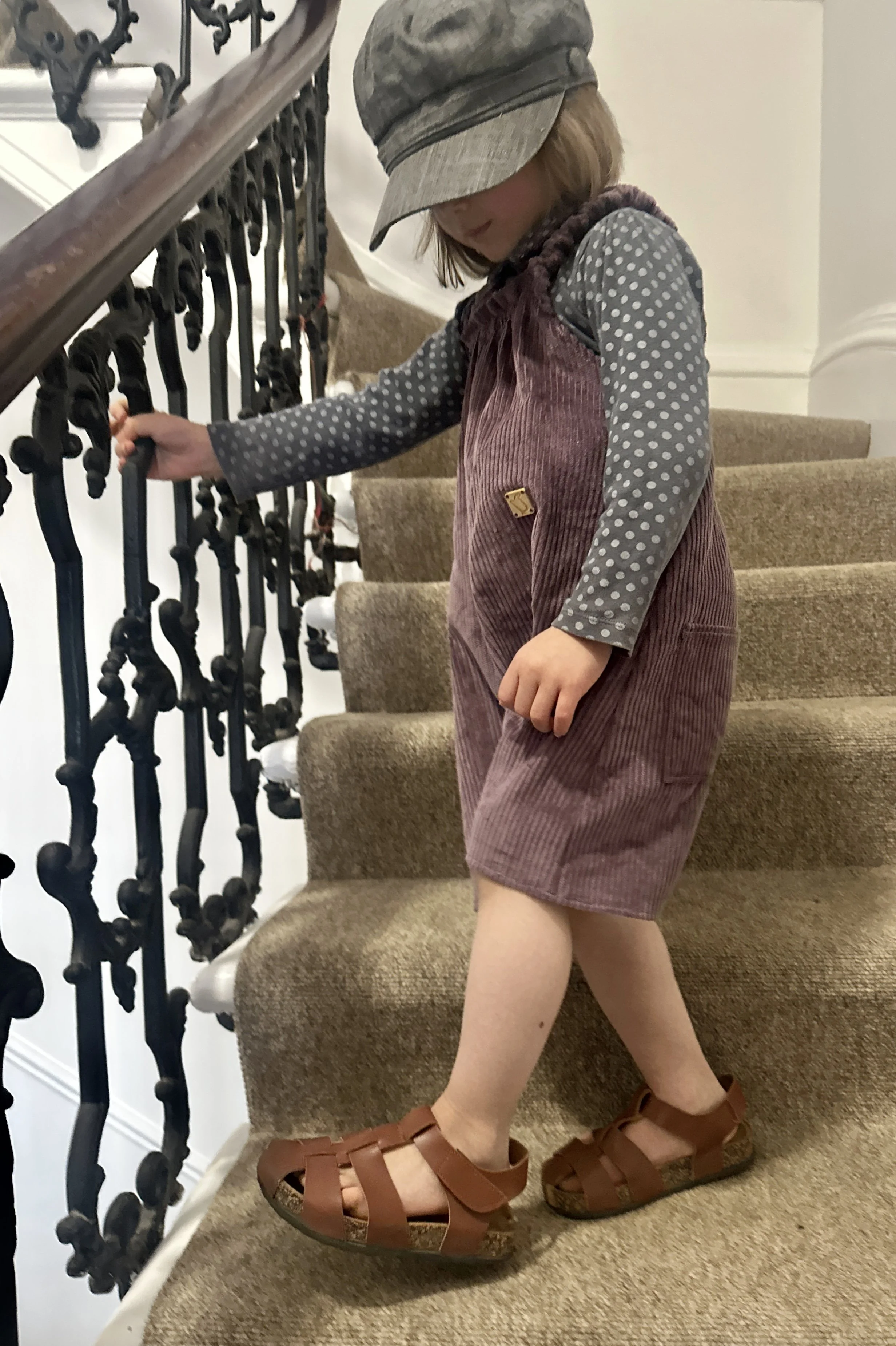 side view of child wearing organic cotton corduroy soft purple short dungarees, layered over a grey dotty top, paired with brown sandals, grey hat