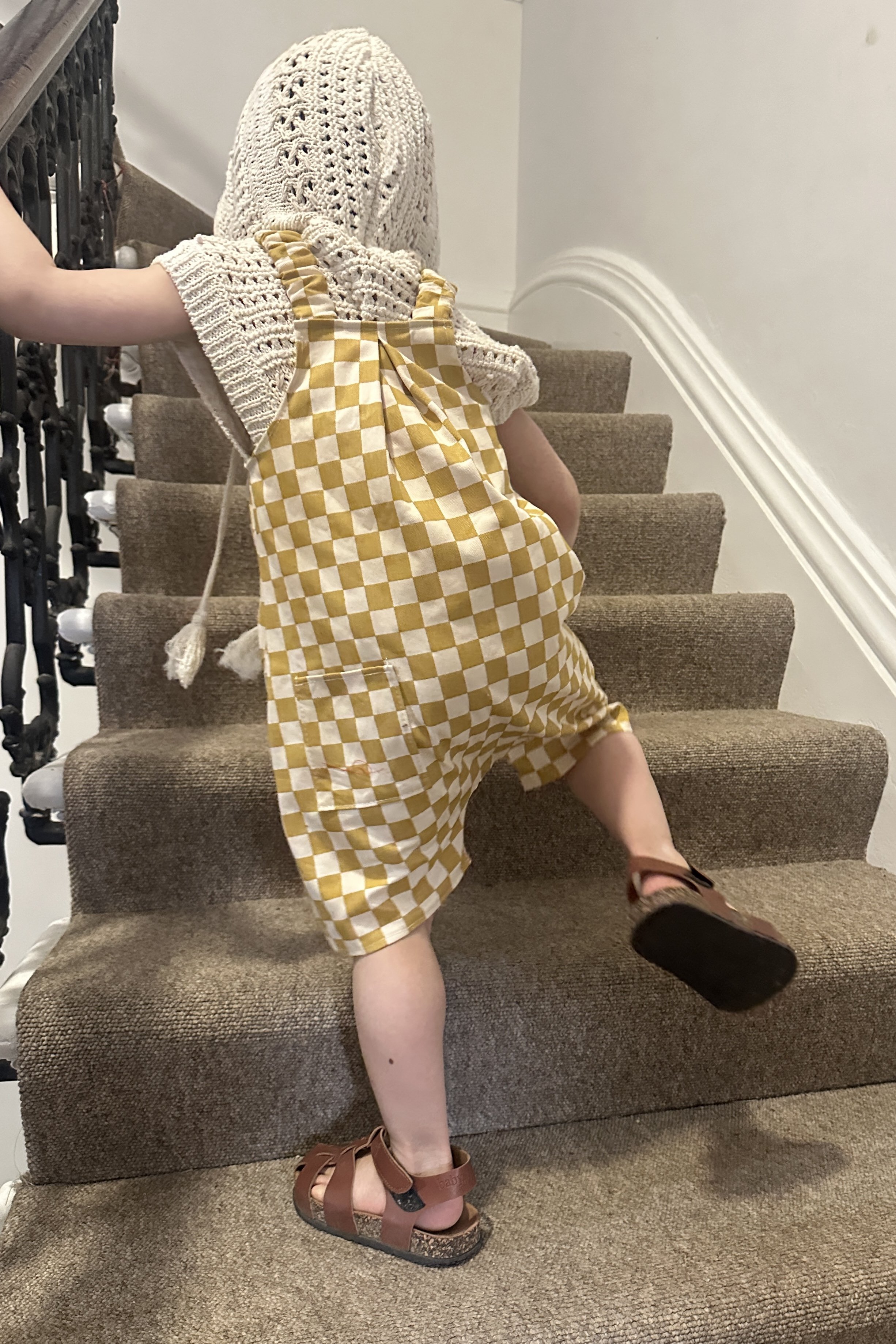 back of child wearing organic cotton canvas pullover short dungarees in mustard checkerboard colour, layered over a crochet short sleeves hoodie top, brown sandals