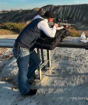 Man shooting a rifle at an outdoor range with mountains in the background.