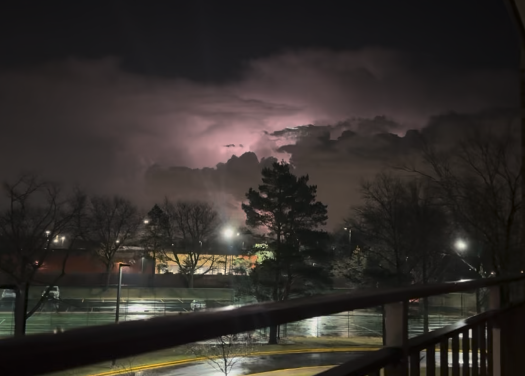 Nighttime scene with dark clouds and lightning in the sky, visible from a balcony overlooking a parking lot and tennis courts with bare trees, illuminated by streetlights.