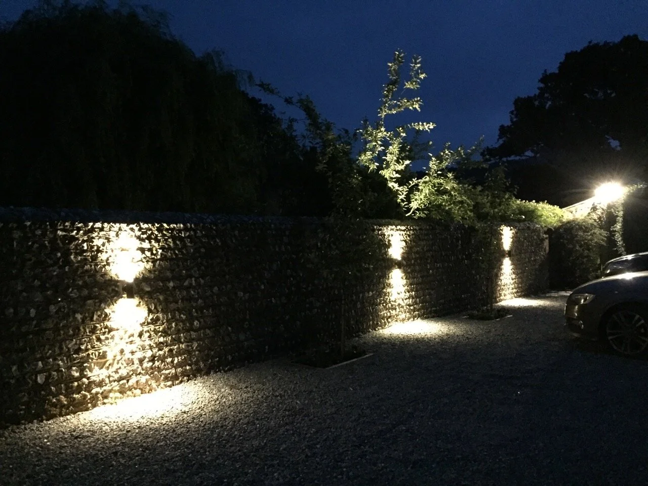 Nighttime outdoor scene with a stone wall illuminated by small lights, trees behind the wall, and a parked car on the right.