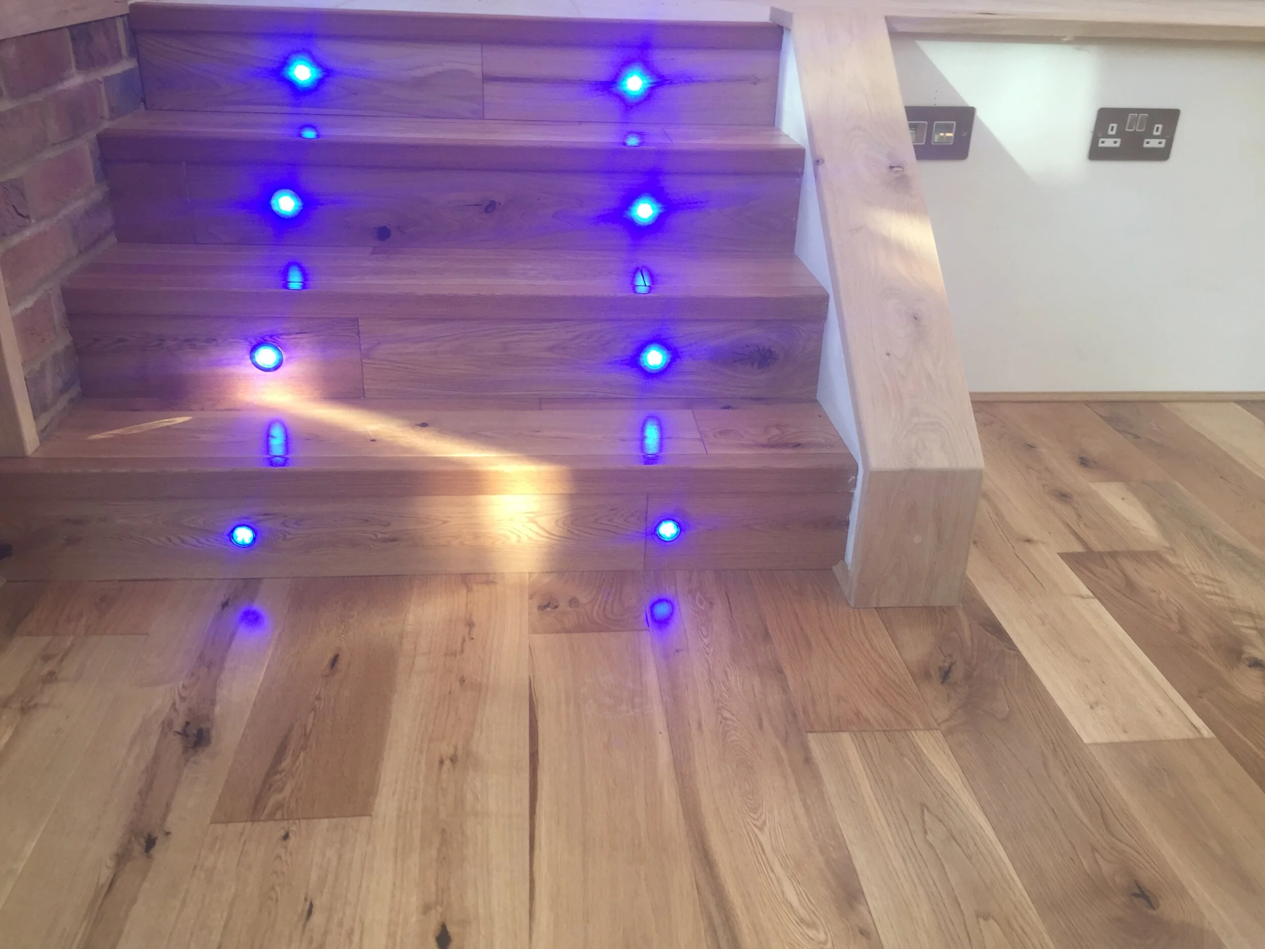 Wooden staircase with blue LED lights embedded in the steps, adjacent to a brick wall and electrical outlets.