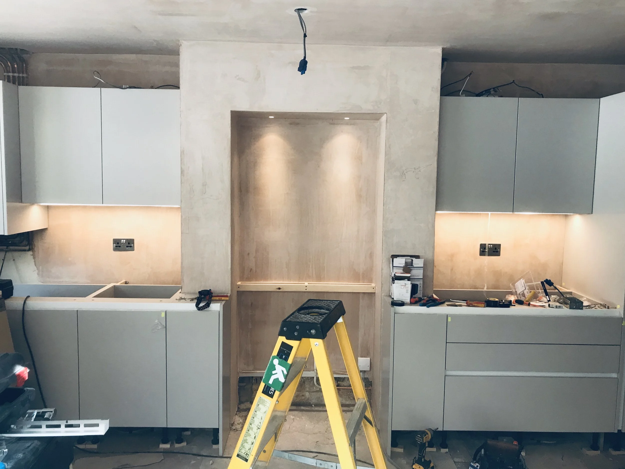 Modern kitchen under construction with gray cabinets, illuminated backsplash, unfinished ceiling wiring, tools, and a yellow step ladder