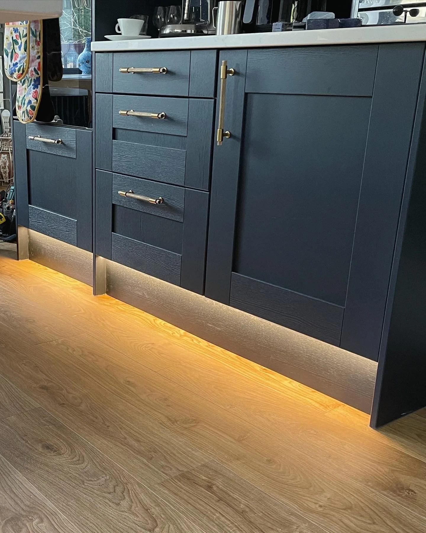 Close-up of a modern kitchen cabinet with dark blue wood finish, gold handles, and under-cabinet lighting on a wooden floor.