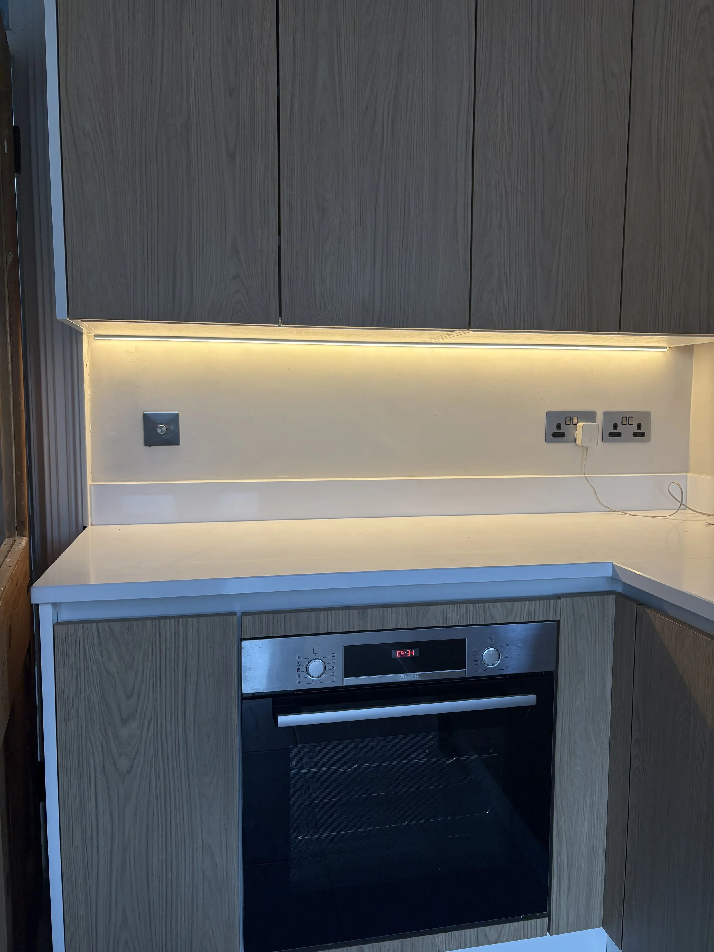 A modern kitchen with a white countertop, wooden cabinets, a built-in oven, and electrical outlets on the wall.