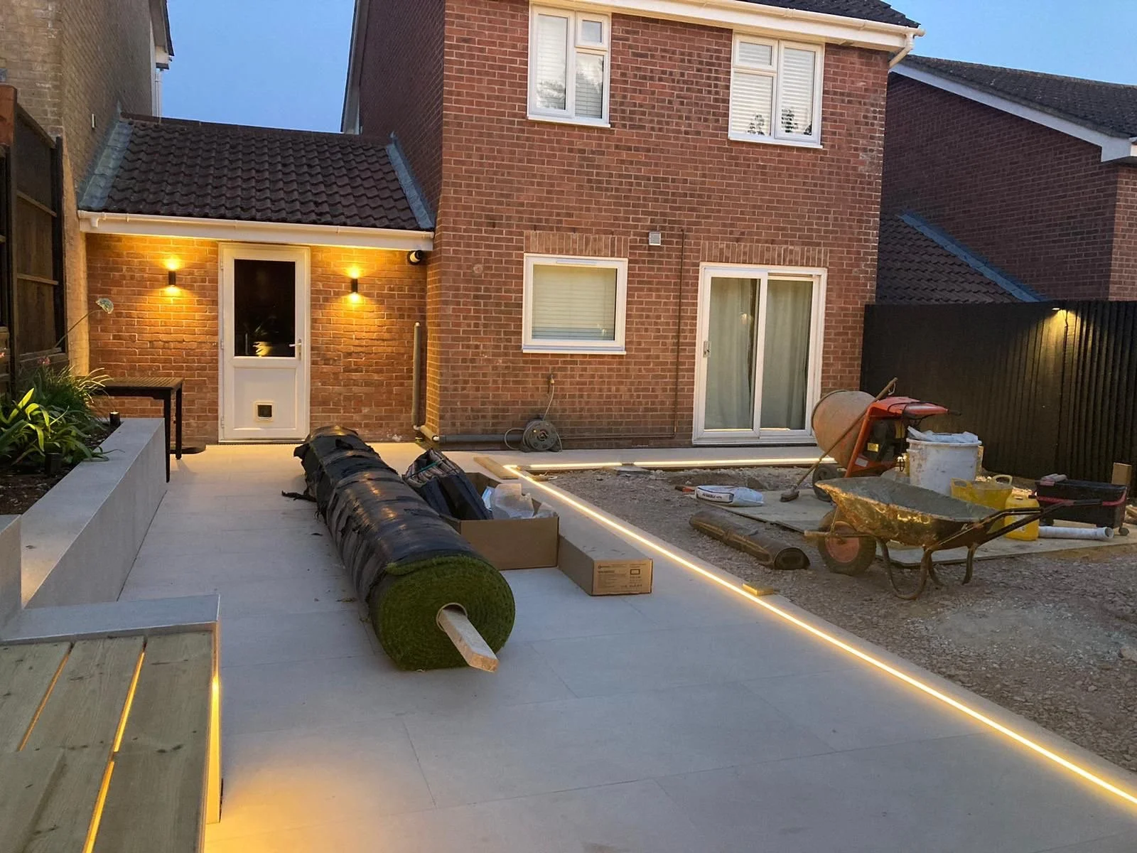 Backyard under construction with building materials, tools, wheelbarrow, and supplies near a brick house with lit outdoor lights.