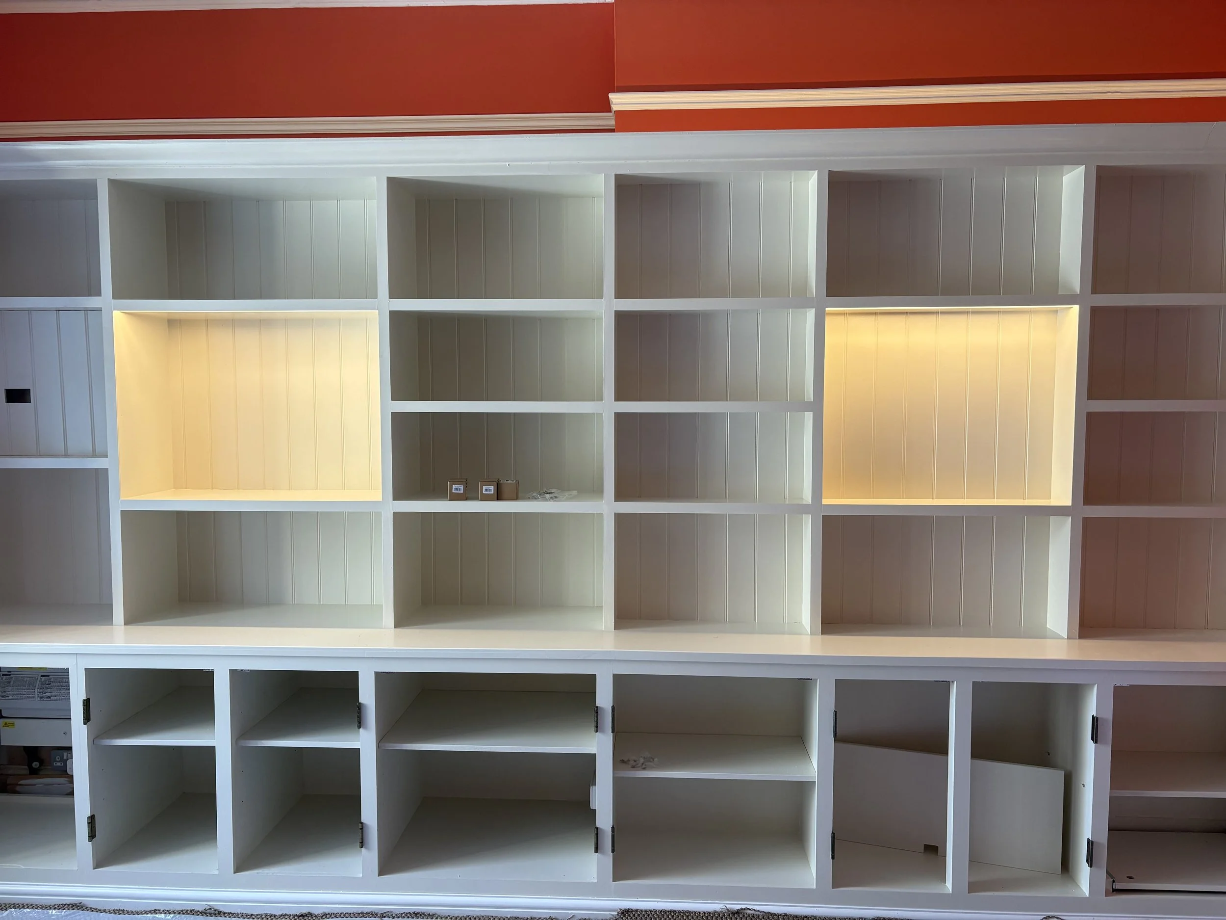 White open-shelving unit with multiple compartments, some illuminated, against a red wall.