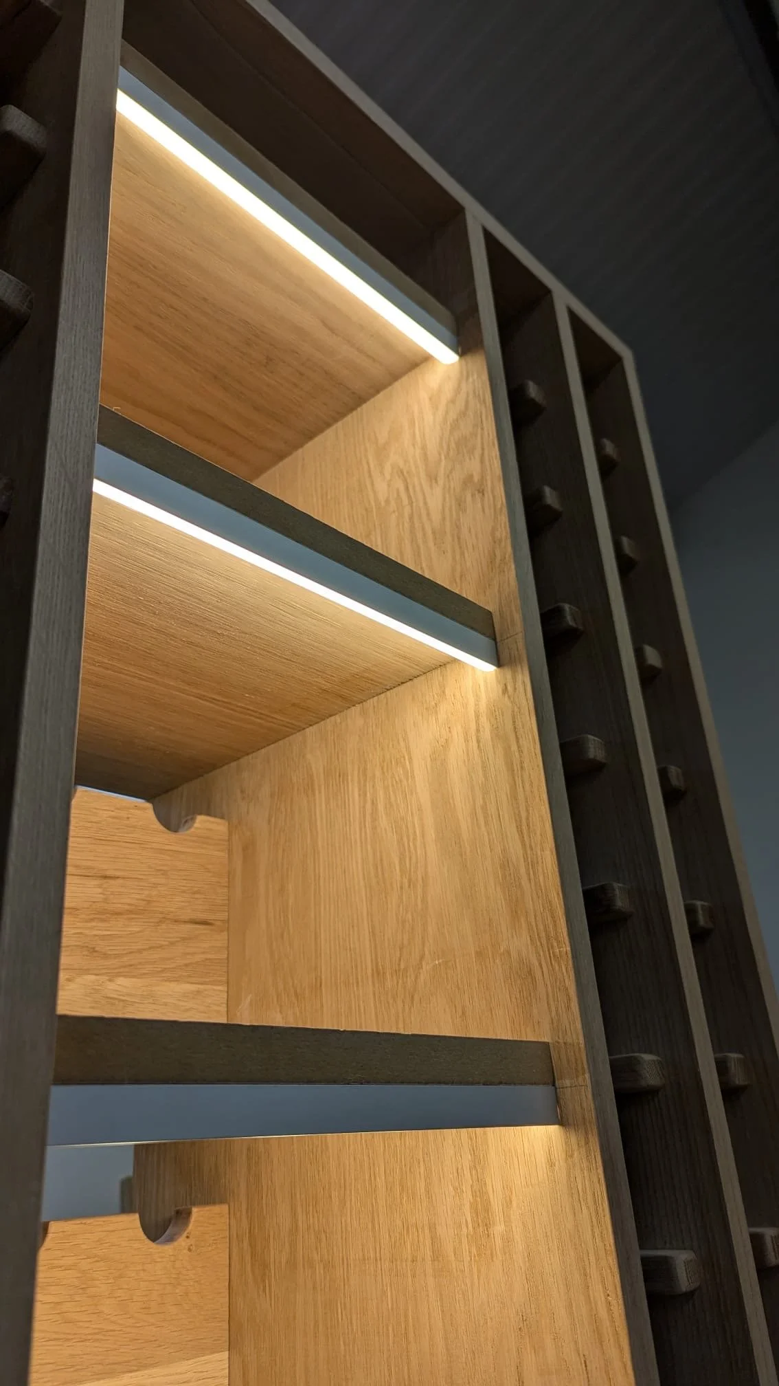 A close-up photo of a wooden wine rack with empty compartments, showing horizontal shelves with a light wood finish and vertical dark wood dividers, lit from above.
