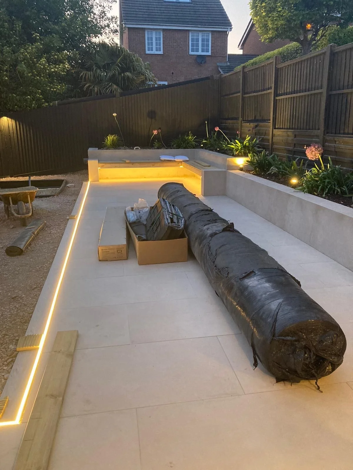 A backyard patio under construction with a large black pipe, wooden planks, a cardboard box, and an LED strip light installed along the edge of the patio.