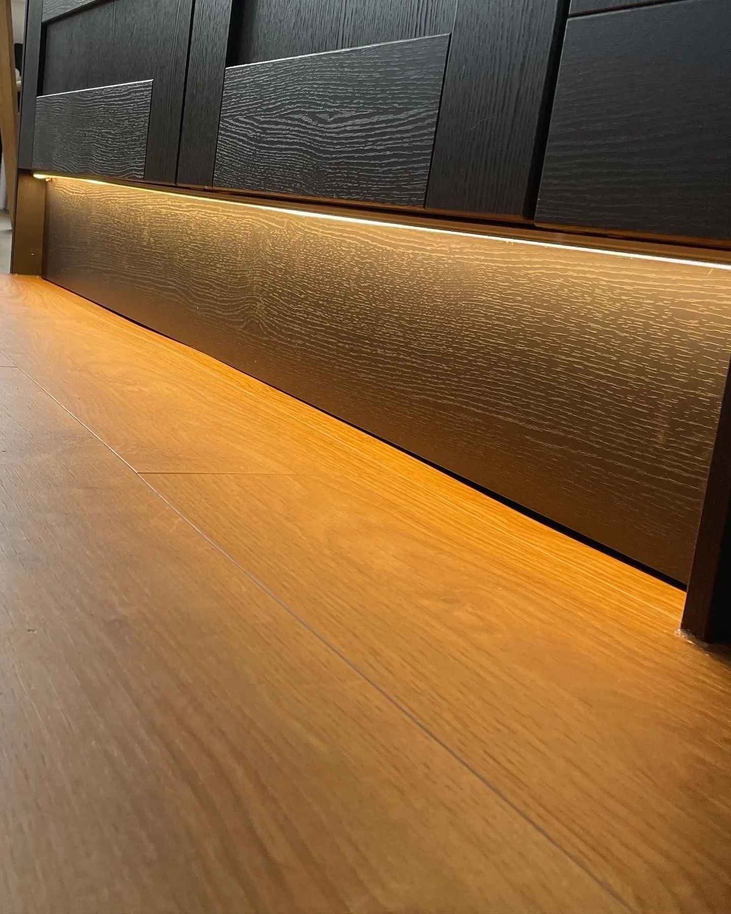 Close-up of a modern black wooden cabinet with a warm illuminated bottom panel, and a wooden floor.