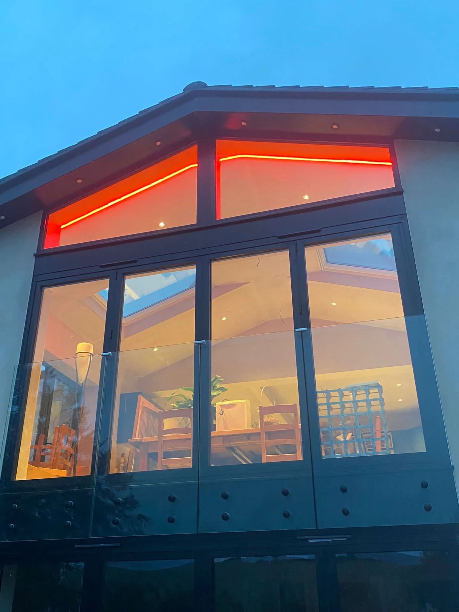 A modern house's upper balcony with large glass windows, red neon lighting, and interior furniture visible inside, captured during twilight.