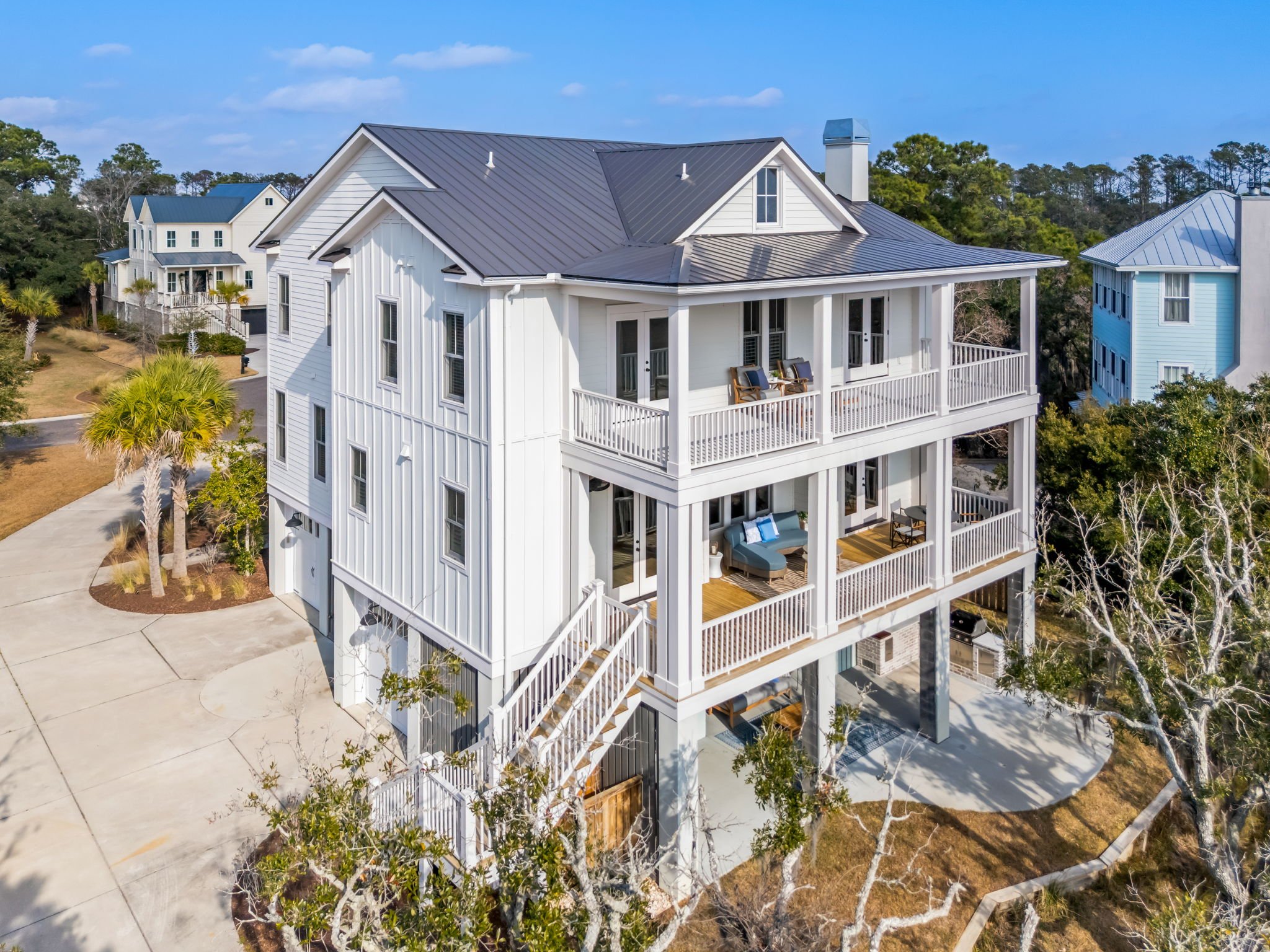 A modern gray house on stilts with a staircase leading to the front door, surrounded by trees and landscaping, under a clear sky.