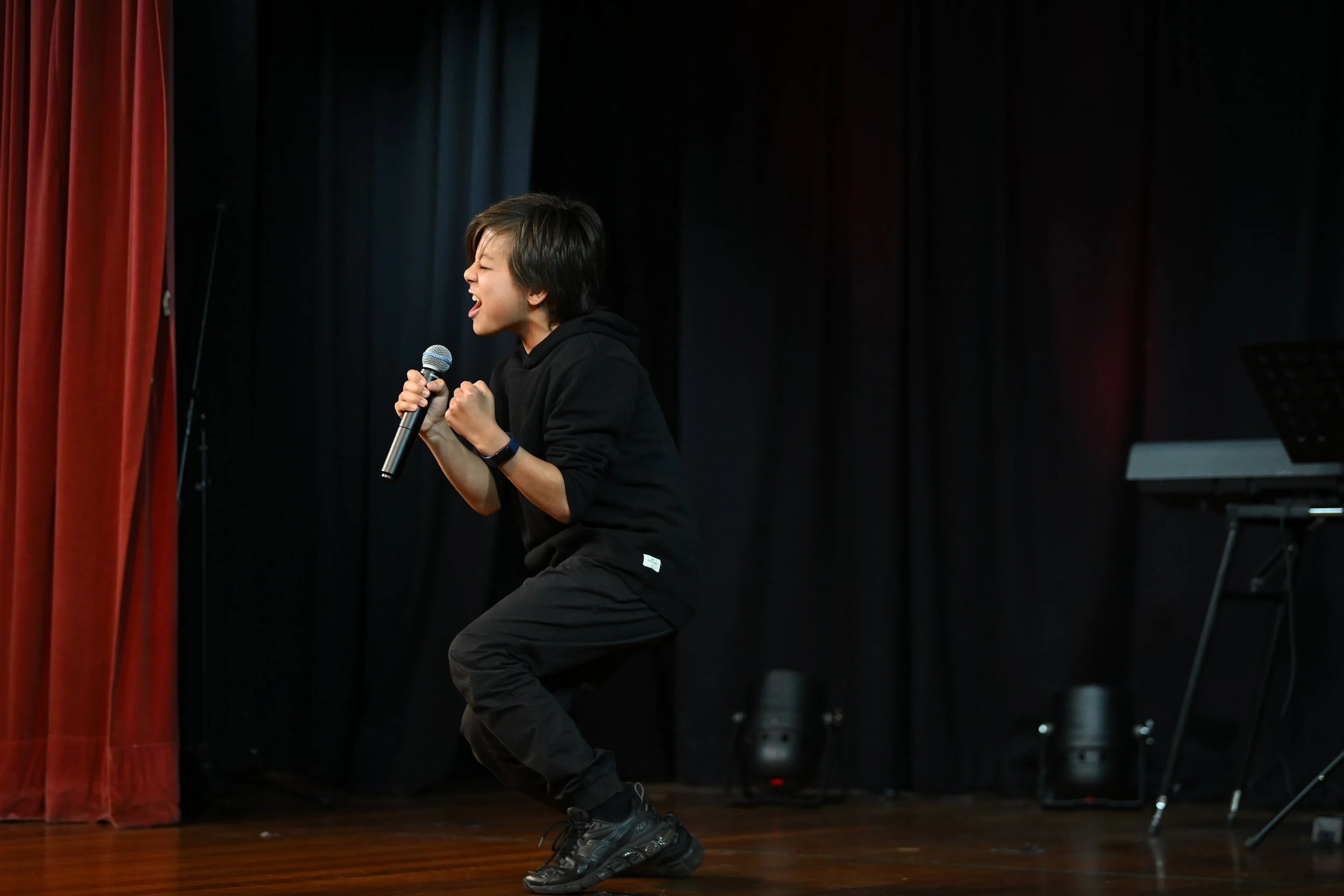 A young person with short dark hair, dressed in black, passionately singing on a stage with a microphone, in front of black curtains with red and blue curtains on the side.