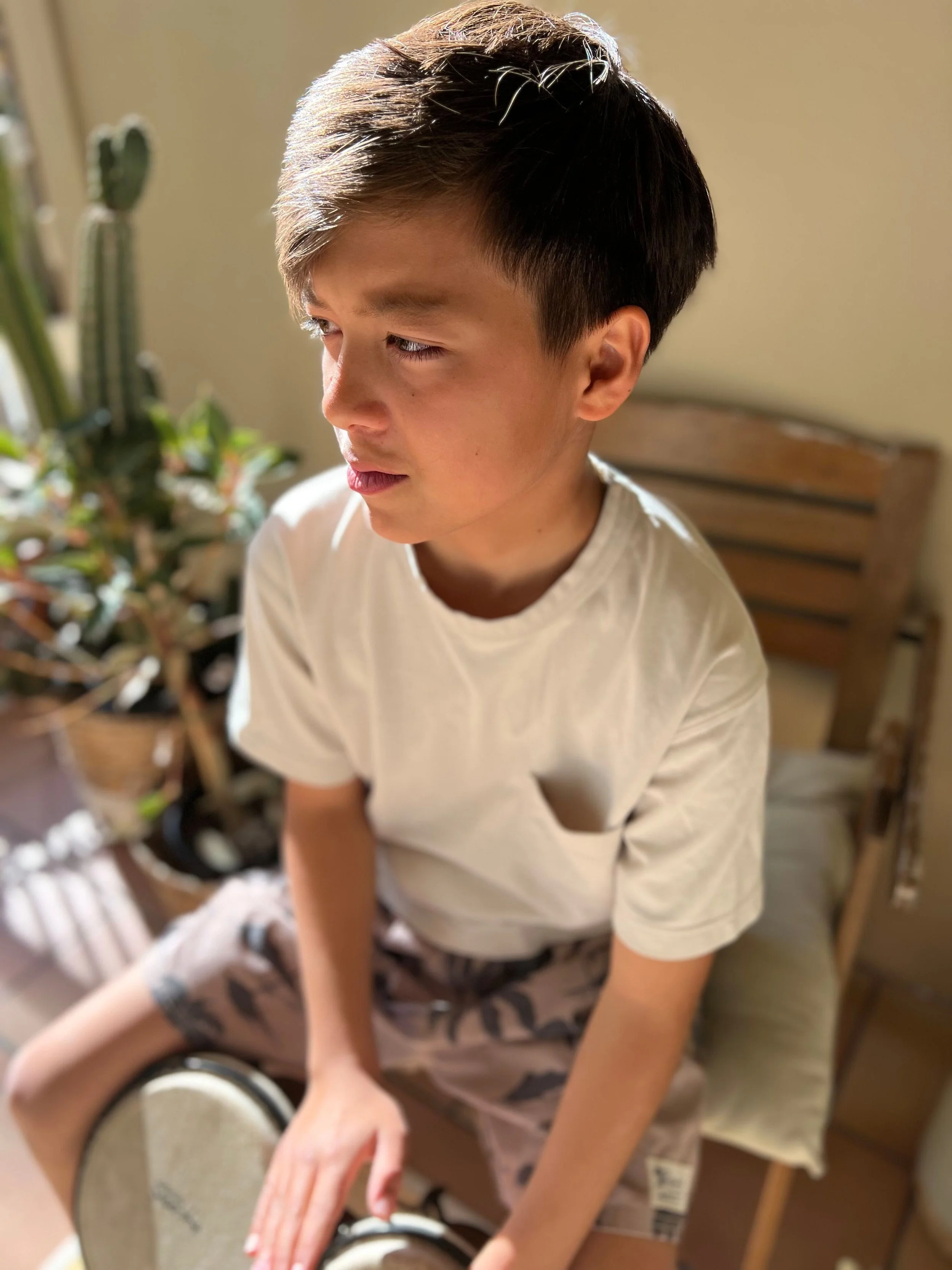 A young boy sitting on a bed, wearing a white t-shirt and camo shorts, with a concerned expression, looking off to the side near a window with sunlight.