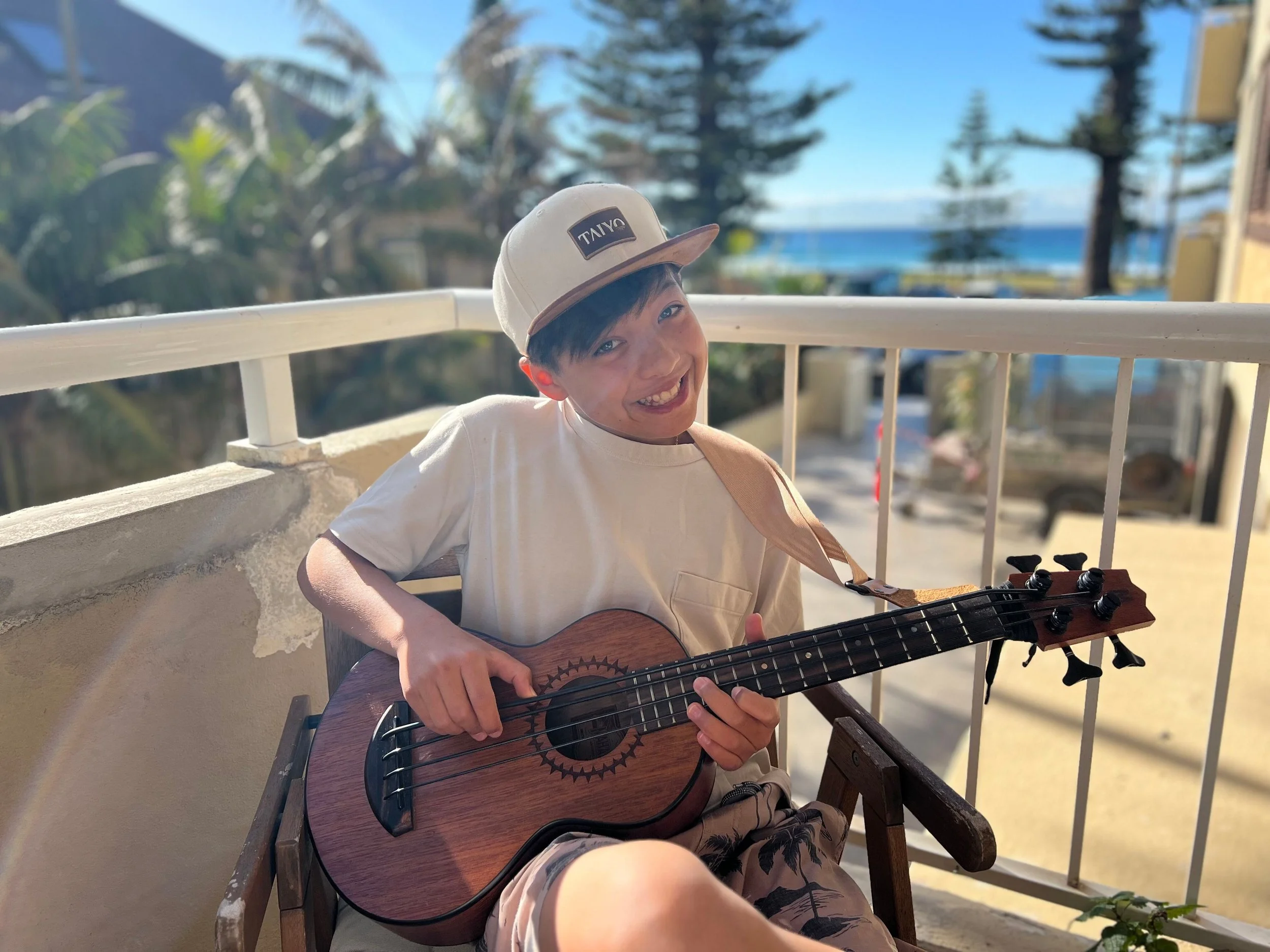 A young boy smiling and playing an acoustic guitar on a balcony with outdoor scenery including trees, a blue sky, and the ocean in the background.