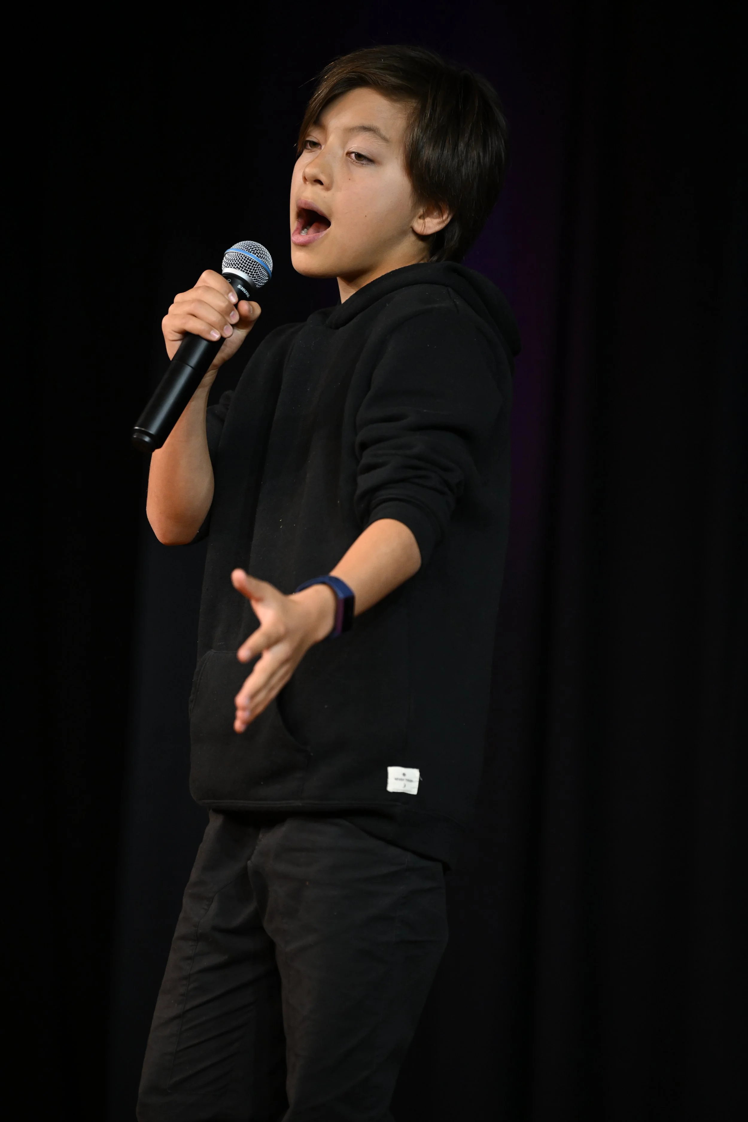 A young person with dark hair, wearing a black hoodie, holding a microphone, speaking or singing on stage with a black curtain background.