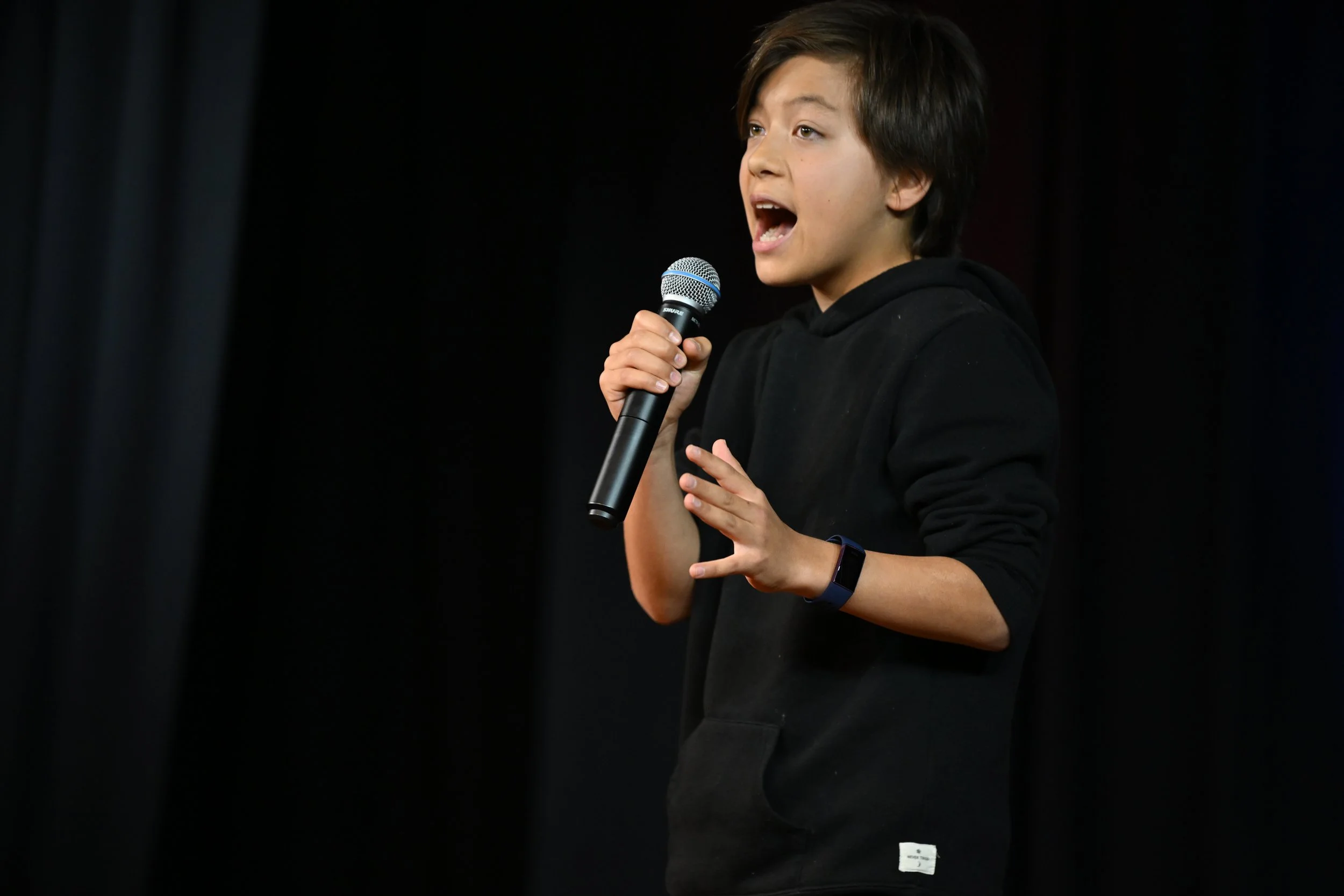 Taiyo Marchand,  A young boy with dark hair wearing a black hoodie holding a microphone, speaking or singing on stage with black curtains behind him.