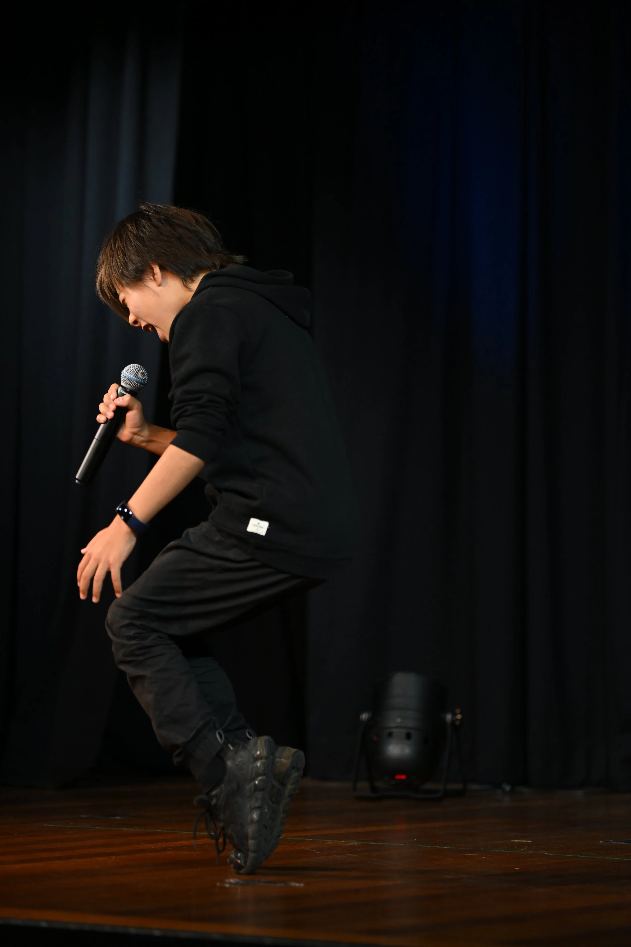 Taiyo Marchand, A person in black clothing holding a microphone, standing on a stage with black curtains in the background.