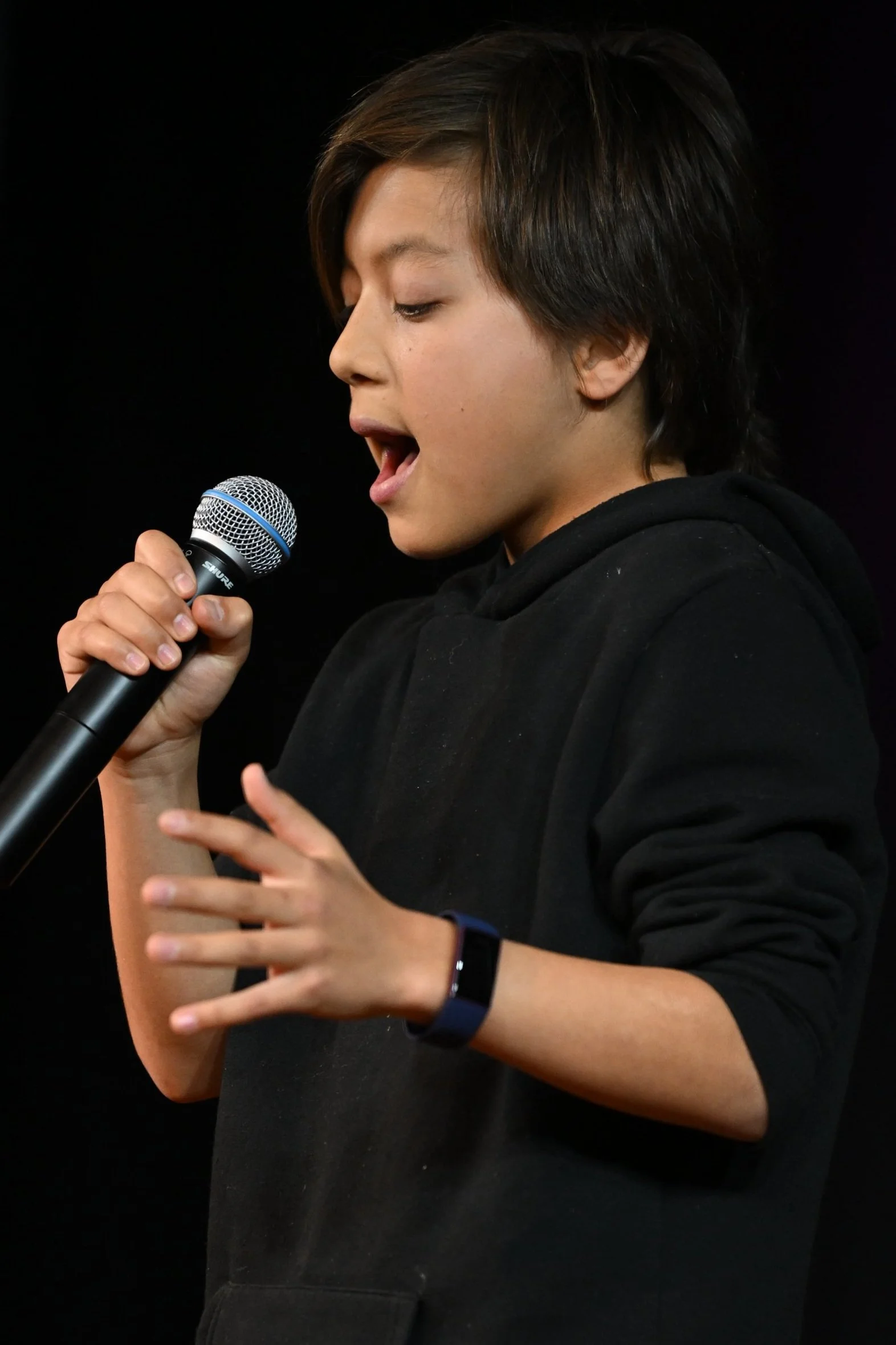 Taiyo, a young boy, singing into a microphone on stage, wearing a black hoodie and a fitness tracker on their wrist, with a black background.