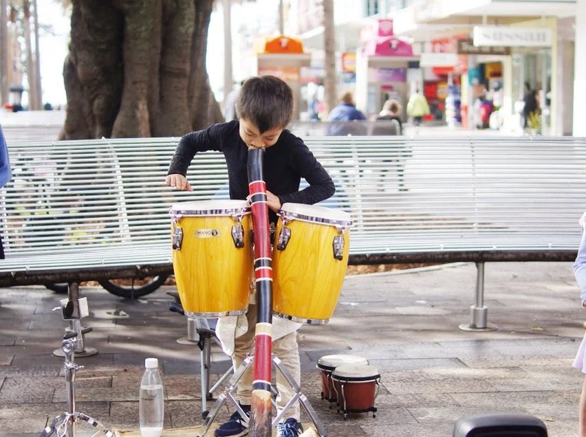 Taiyo playing a large didgeridoo outdoors near a bench and other drums, with a shopping area in the background.