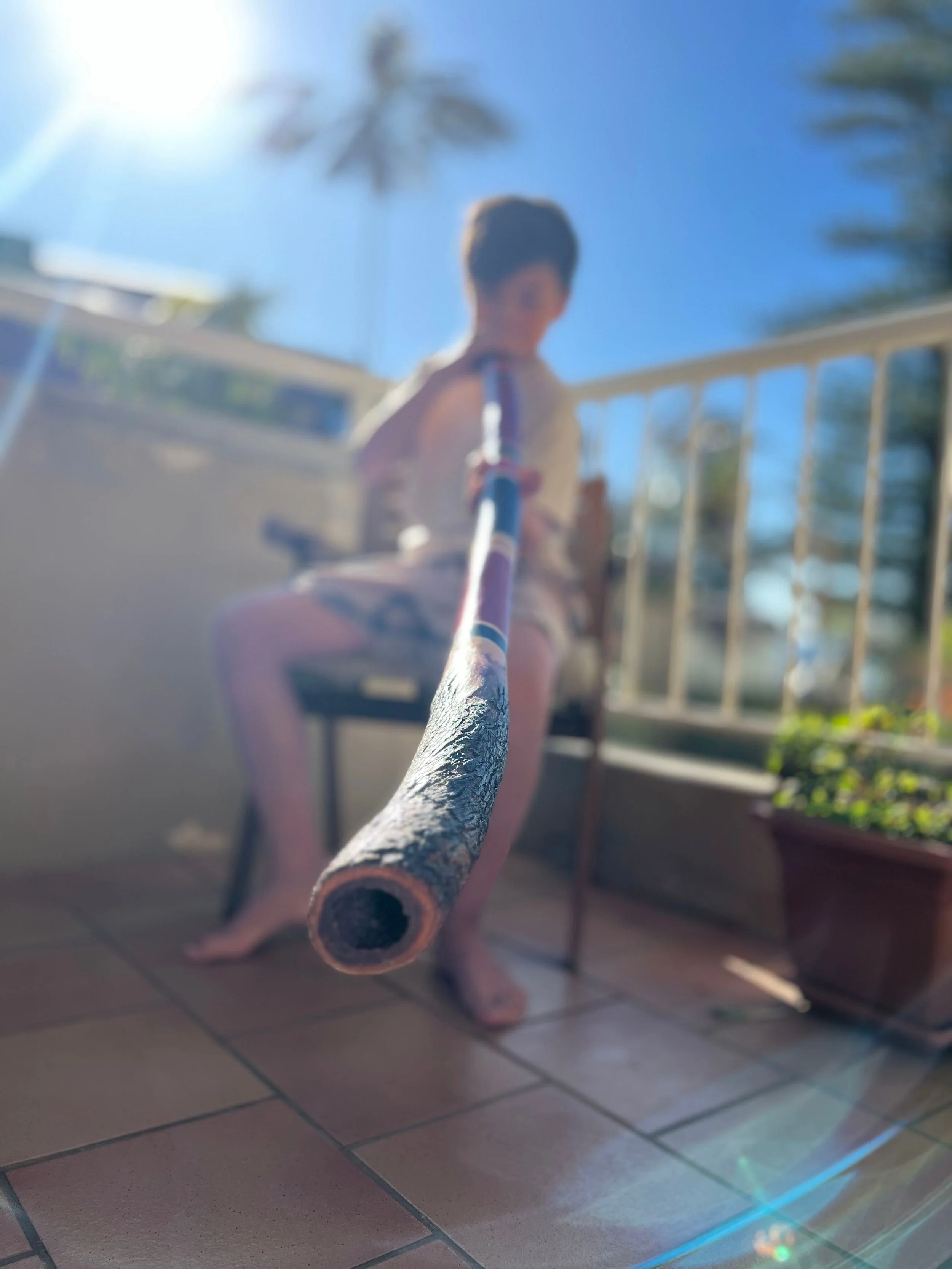 Taiyo Marchand, Child sitting on a chair on a balcony, holding a long stick towards the camera, with bright sunlight and blue sky in the background.