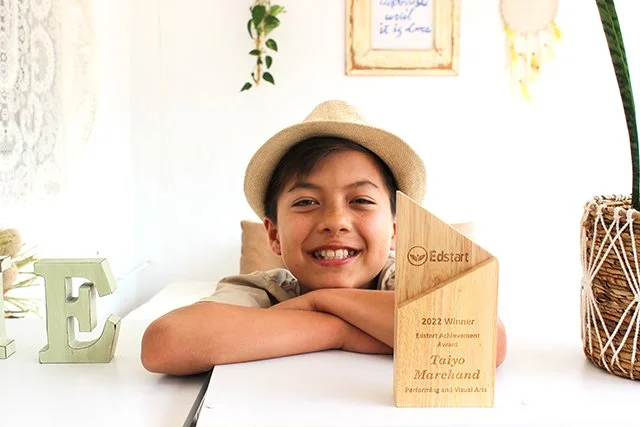 Taiyo Marchand, A young boy with a hat smiling at the camera, sitting at a white table with a wooden award plaque in front of him.
