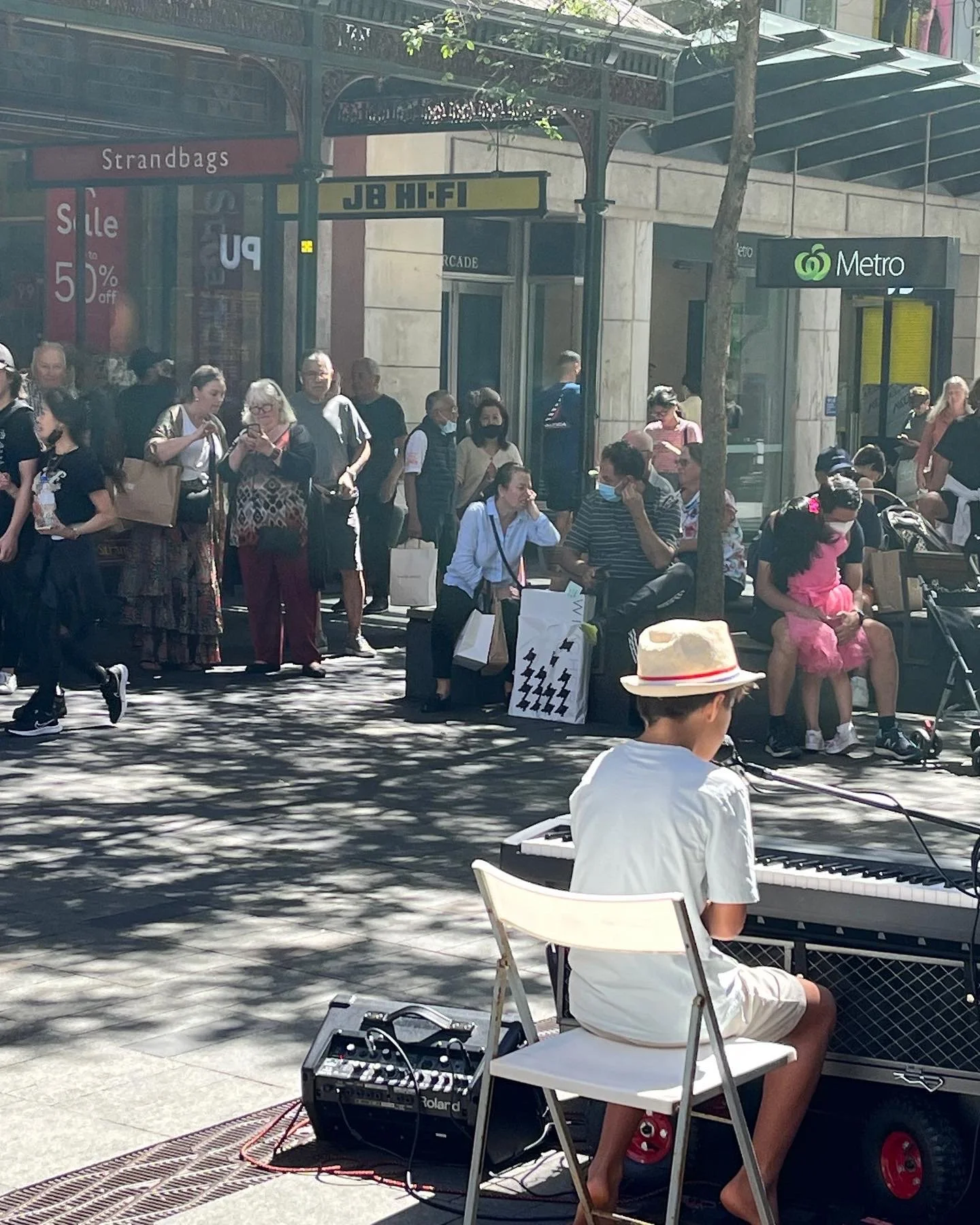 Taiyo, a young boy wearing a white shirt, shorts, and a straw hat playing a keyboard on a sidewalk at a busy city metro station entrance with a crowd of people waiting or walking nearby, some sitting on benches and others standing.