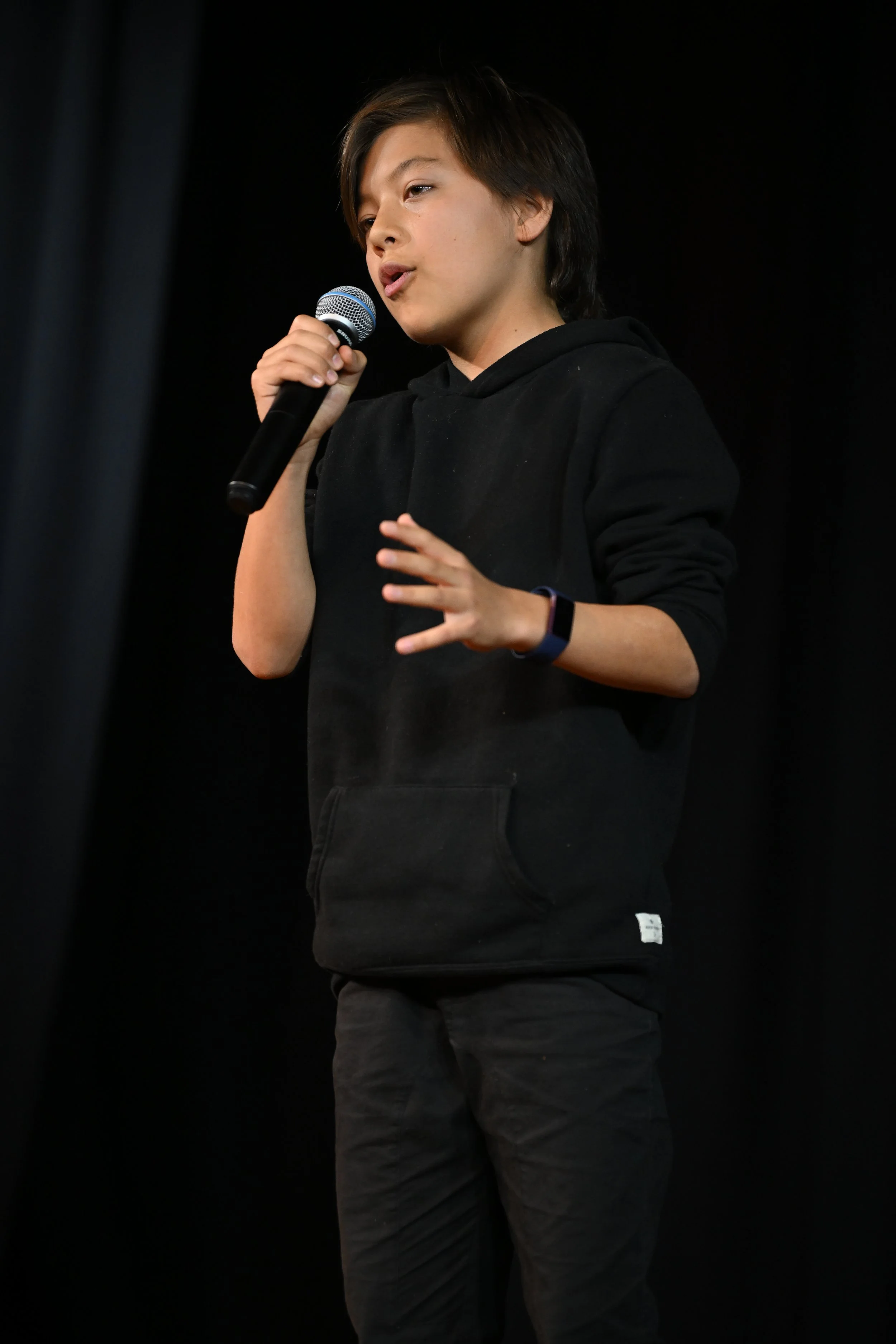 A young person with short dark hair, wearing a black hoodie, holding a microphone, and speaking on stage against a dark background.