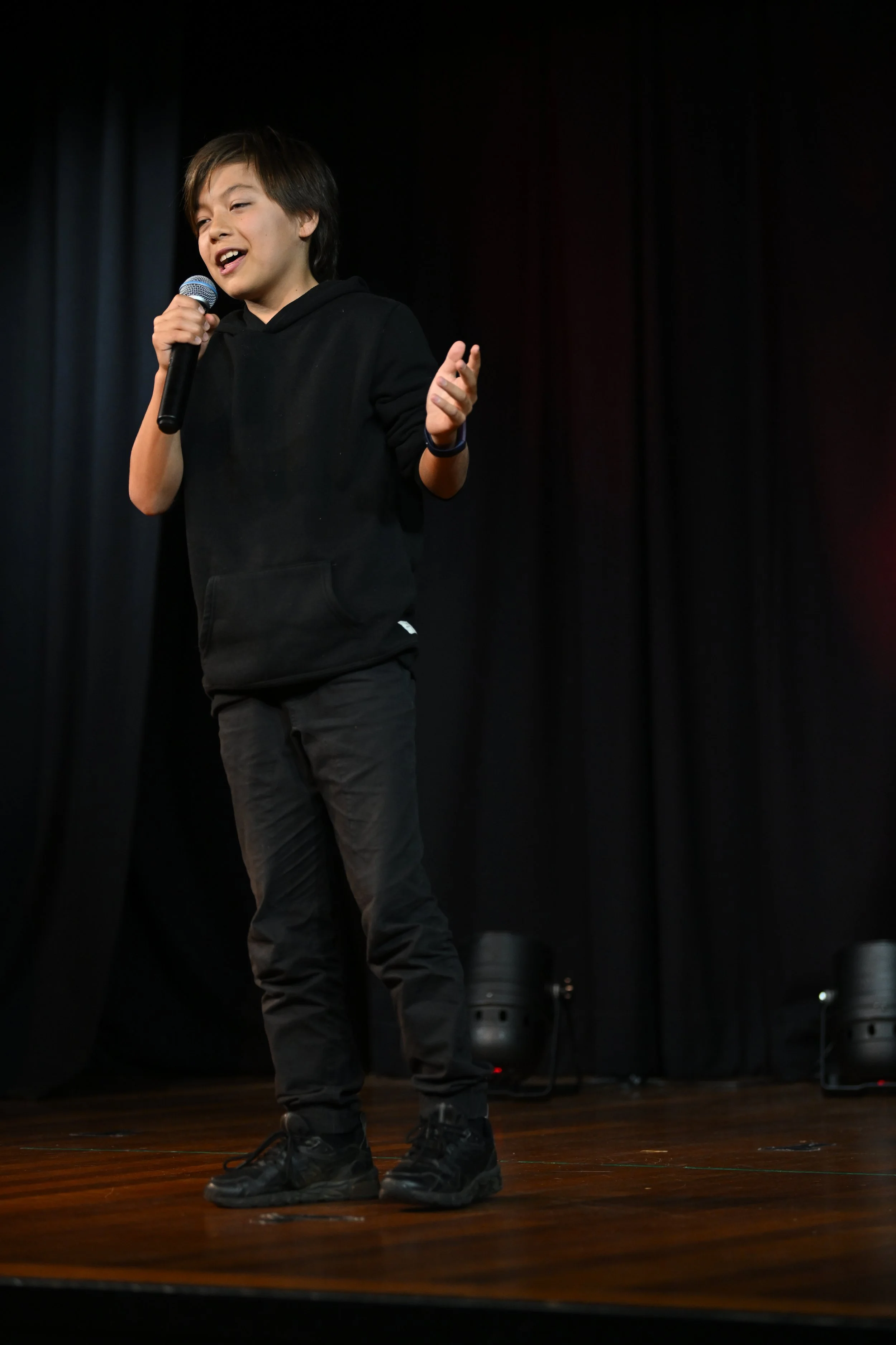 Young boy with dark hair singing into a microphone on stage with black curtains in the background.
