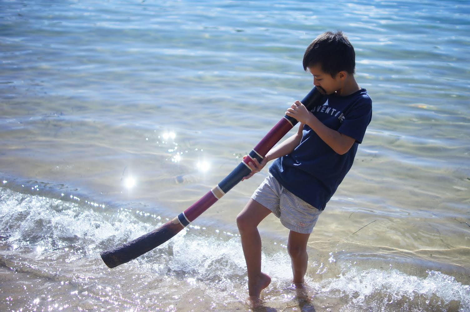Taiyo, a young boy, standing in shallow water at the beach, holding and looking through a long, colorful telescope.