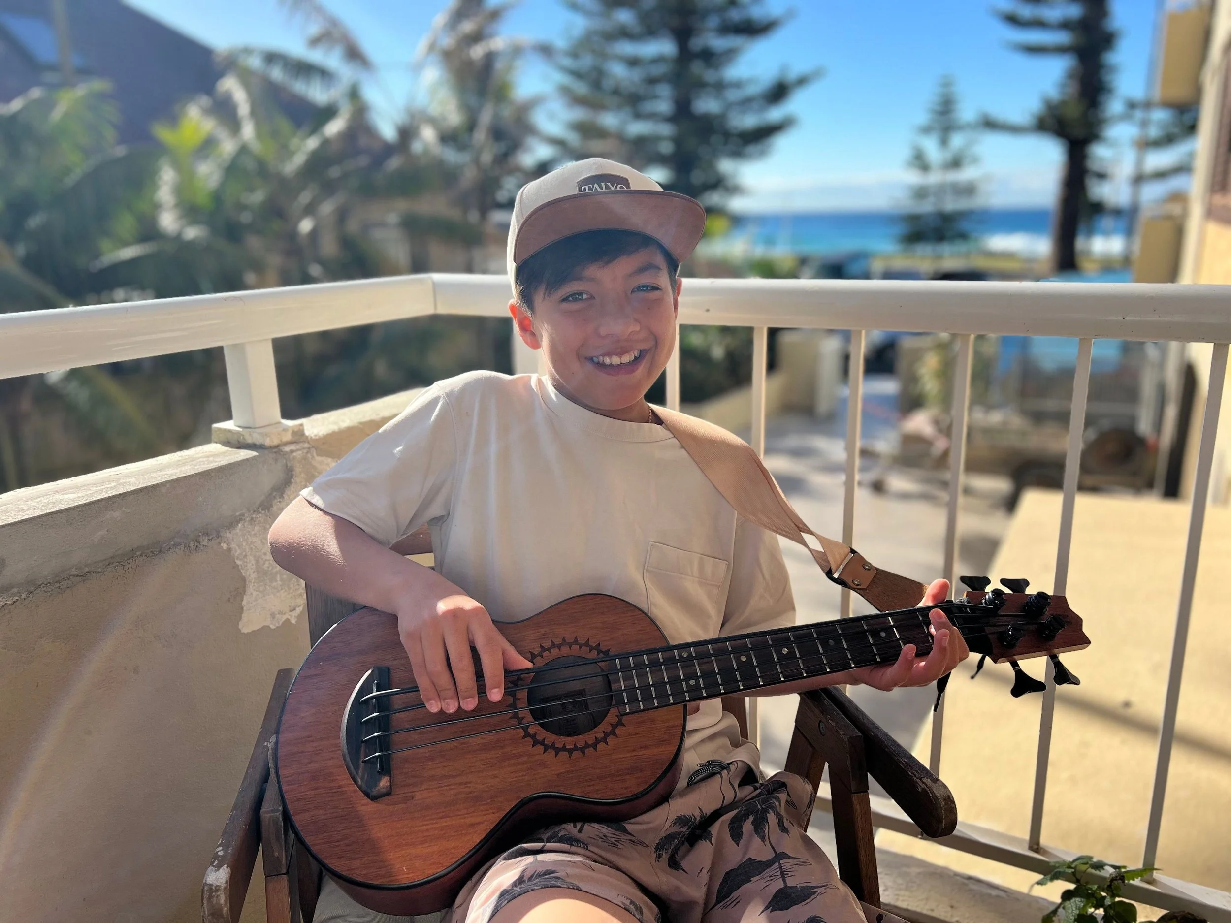 Taiyo Marchand,  A boy sitting on a wooden chair outdoors, smiling, holding an ukulele, with a beach and trees in the background.