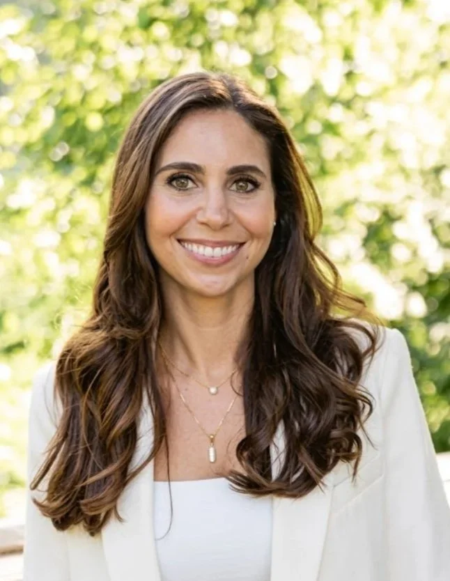 A woman with long, wavy brunette hair smiling outdoors, wearing a white top and blazer, with a blurred green leafy background.