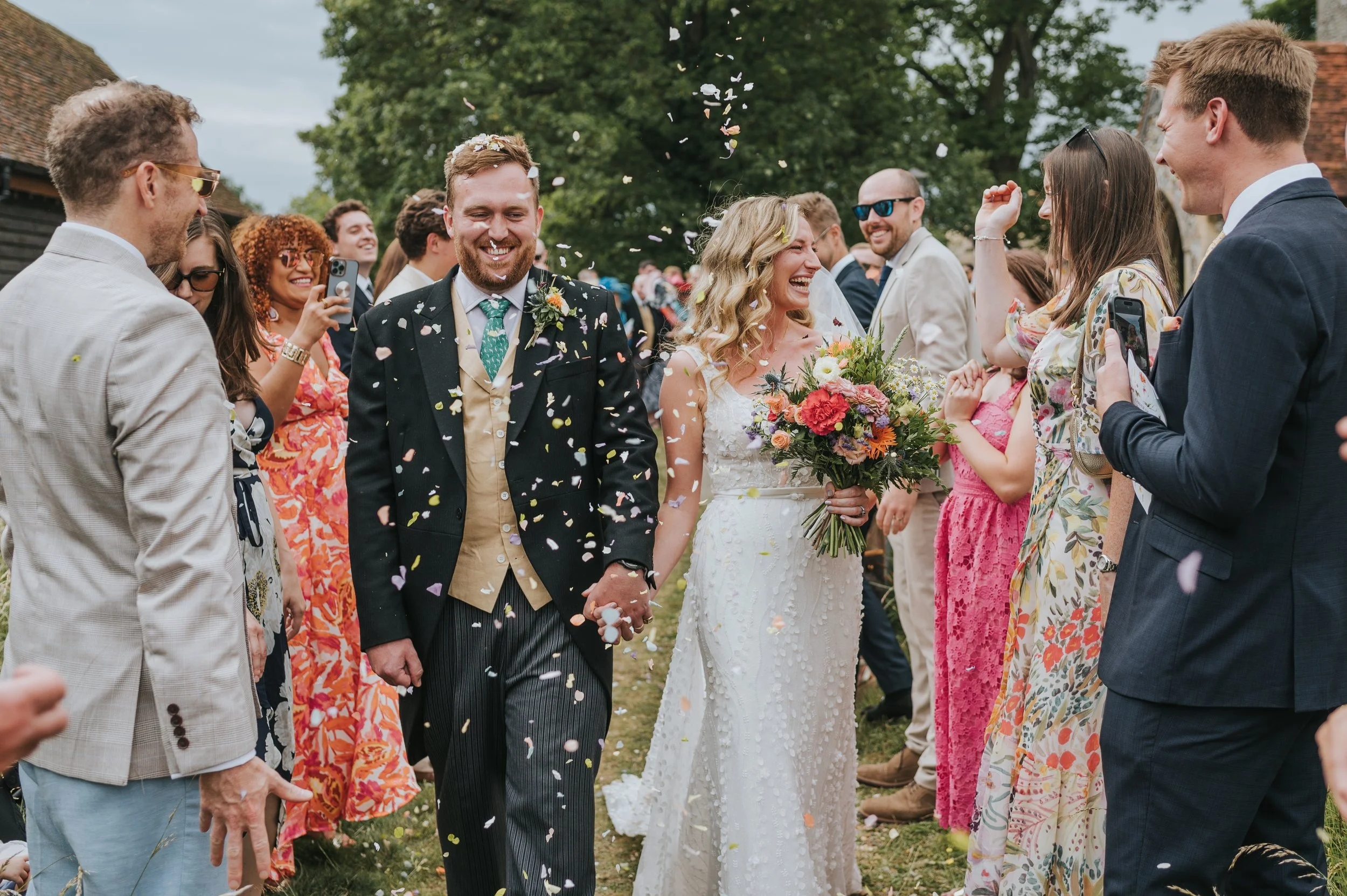 A couple in wedding attire walking hand-in-hand through a celebratory gathering of friends and family, with confetti falling around them, outdoors near green trees.