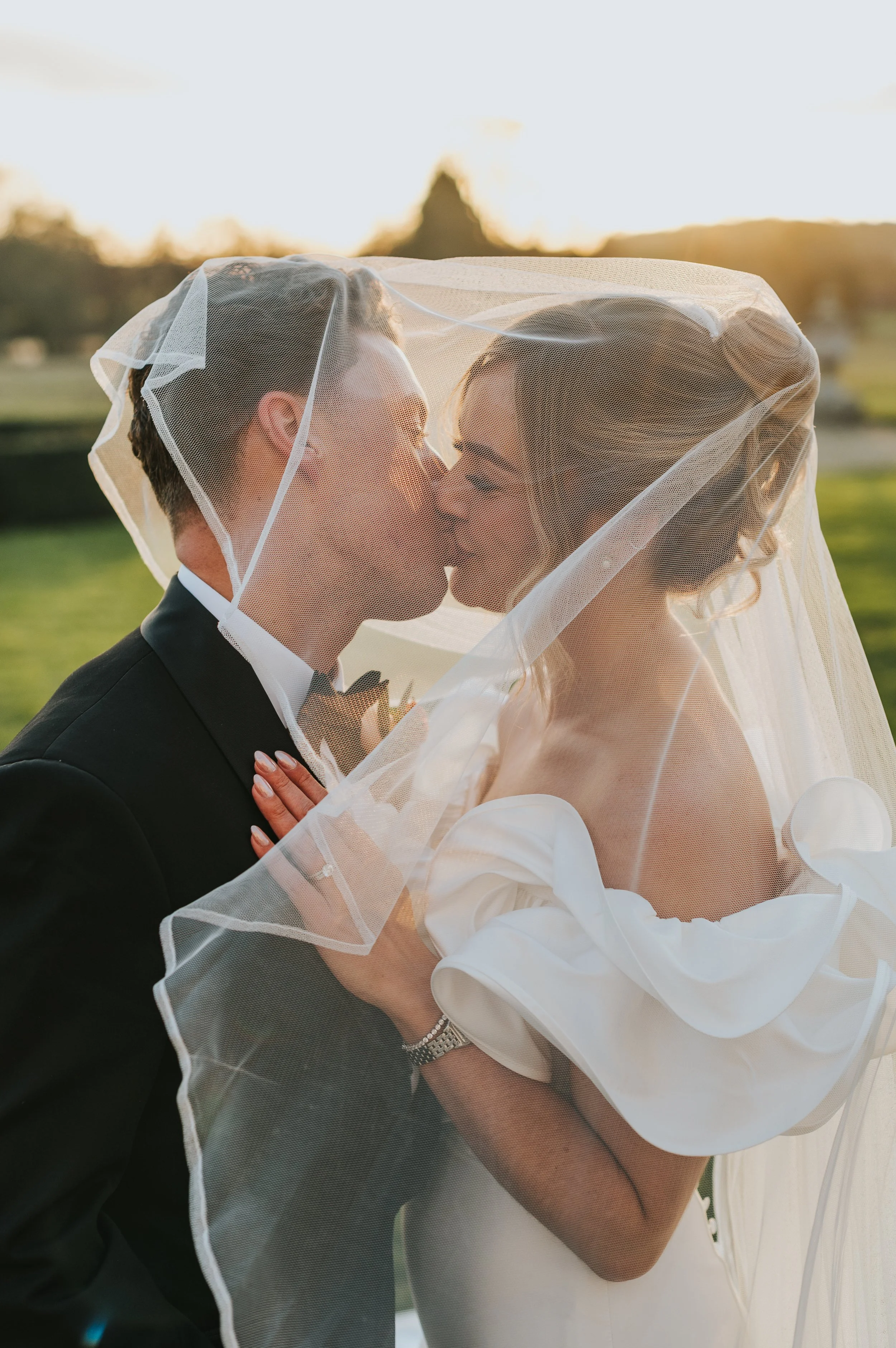A bride and groom sharing a kiss under a veil outdoors at sunset. Rebecca Louise Photography Essex Wedding photographer