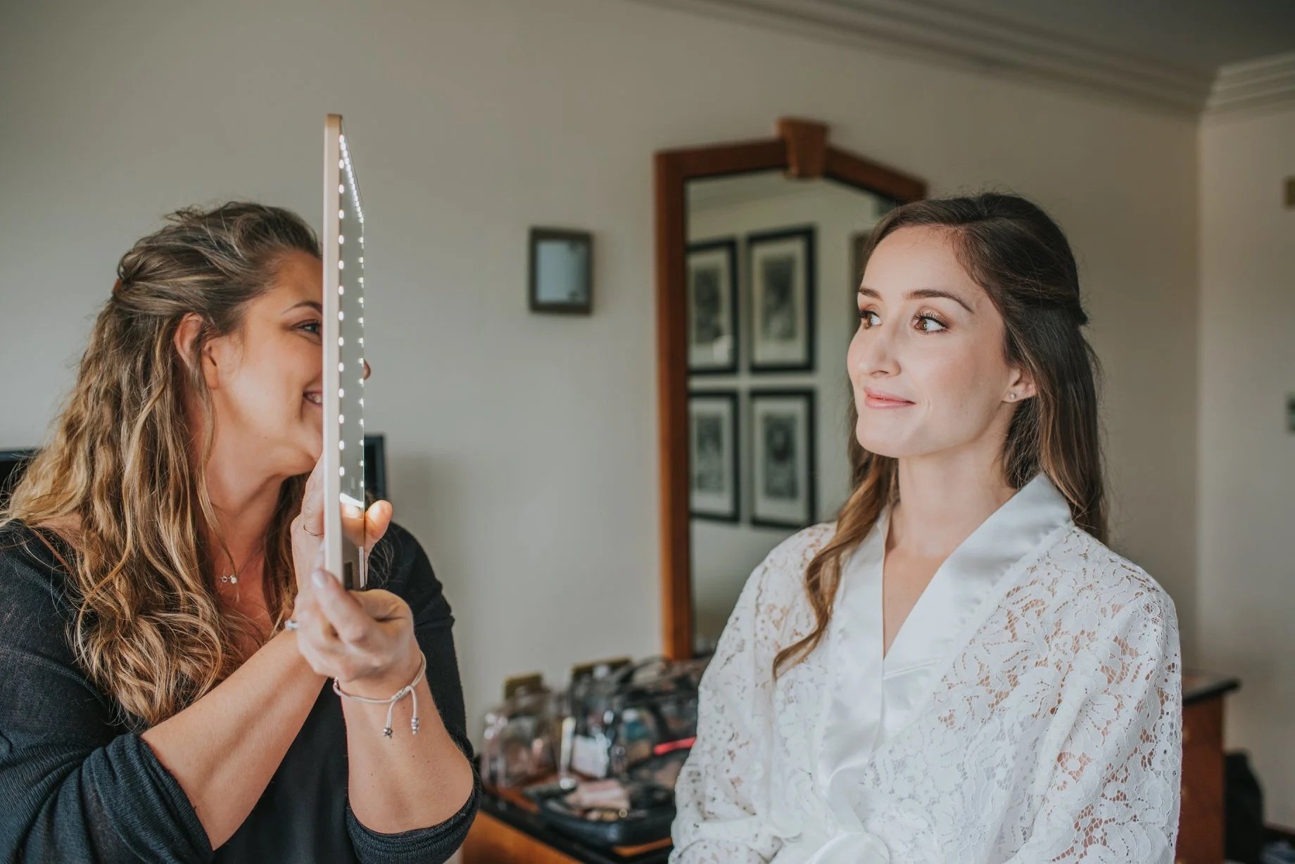A woman is holding a mirror while another woman in a white lace robe looks at her with a slight smile. Rebecca Louise Photography Essex Wedding photographer