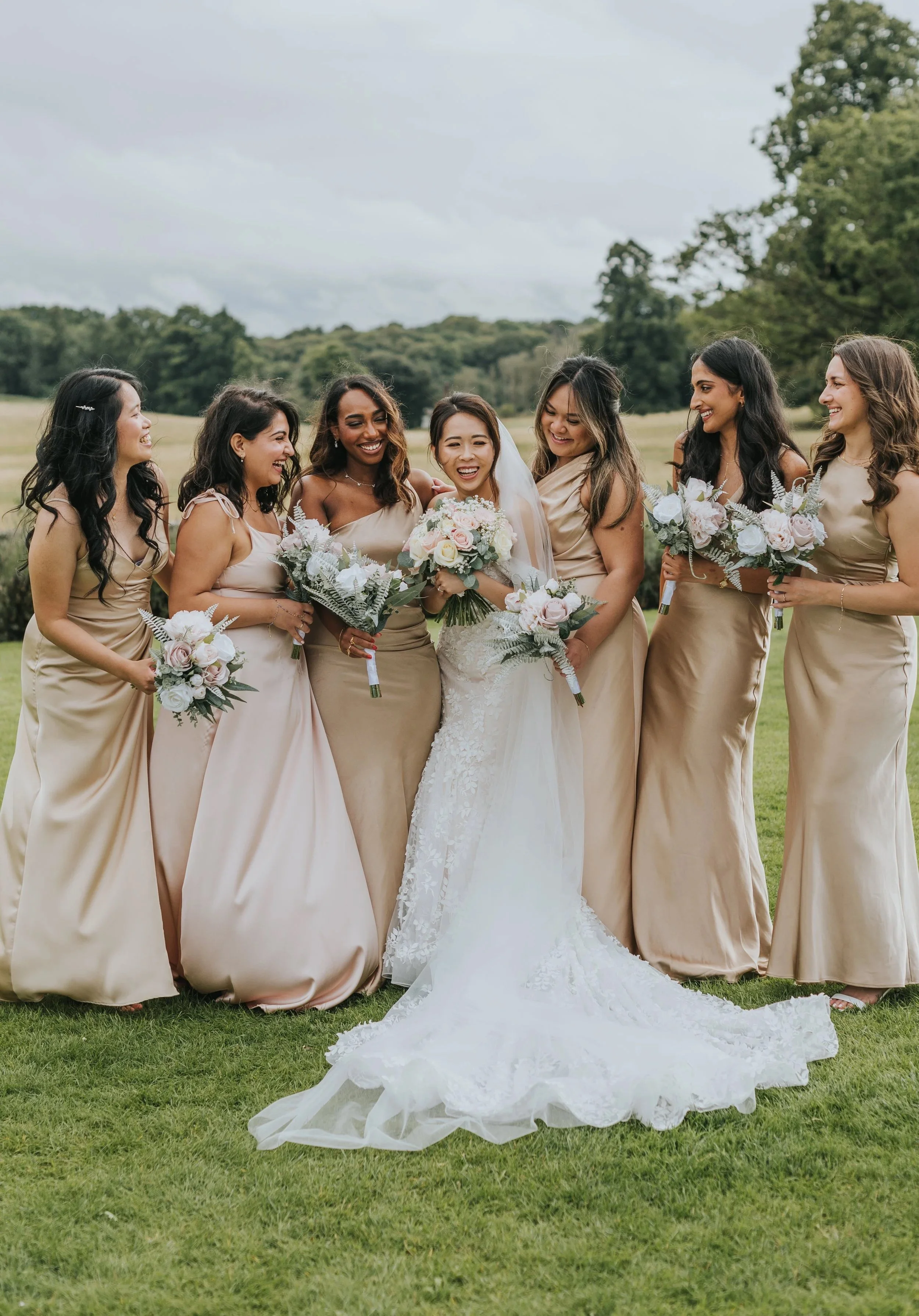 A bride and seven bridesmaids standing outdoors on a grassy field, all smiling and holding bouquets, with trees and a cloudy sky in the background.
