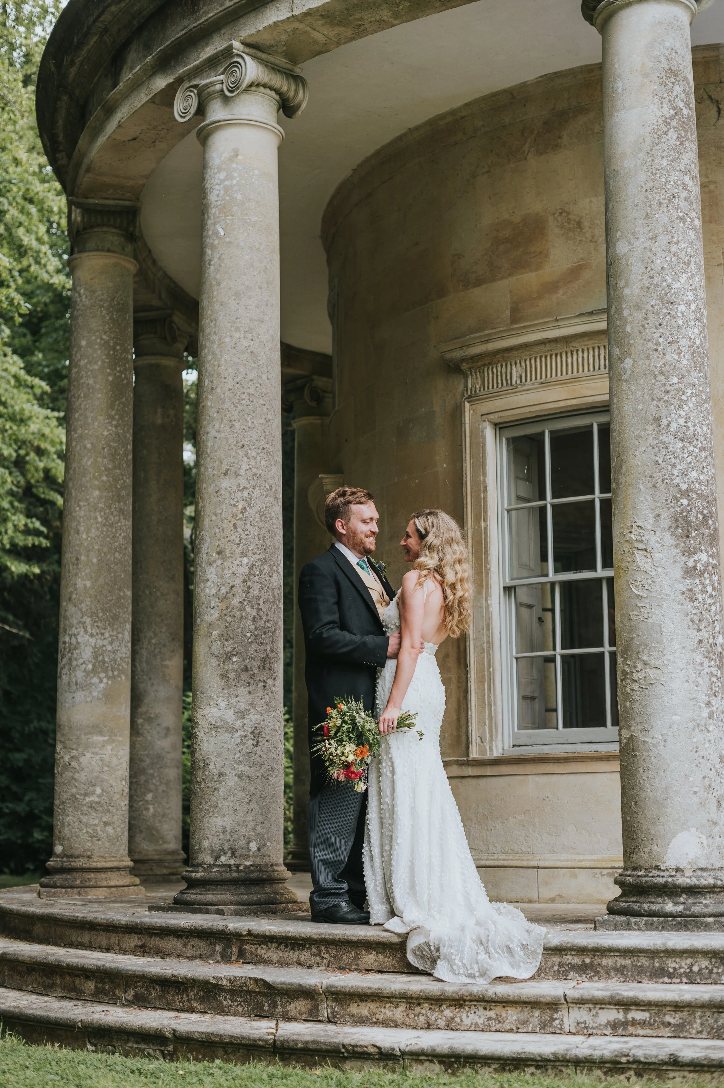 A bride and groom standing on stone steps in front of an old building with tall columns, sharing a moment on their wedding day. Rebecca Louise Photography Essex Wedding photographer