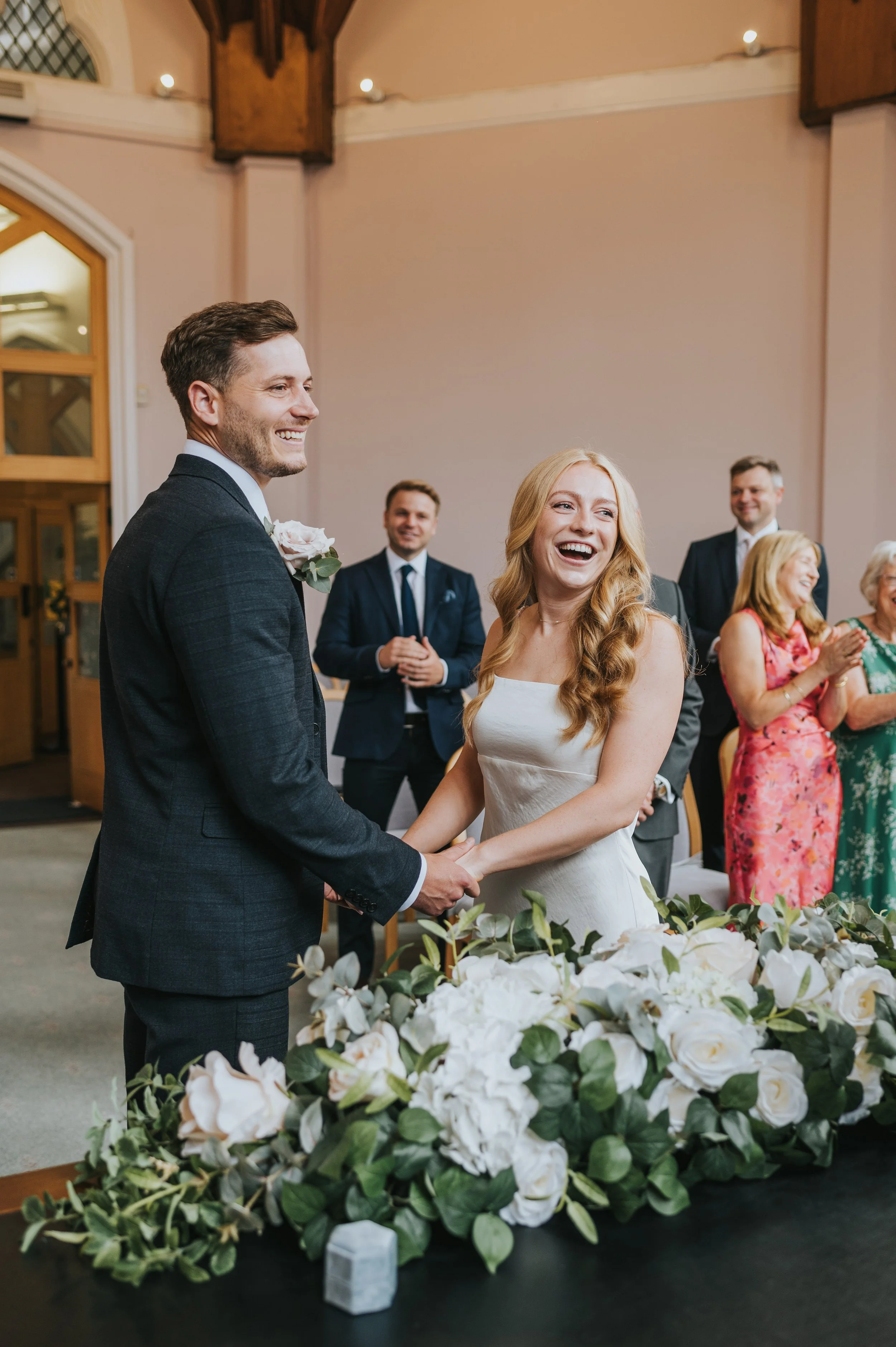 A bride and groom holding hands and smiling during their wedding ceremony, surrounded by friends and family in a warmly lit indoor venue decorated with white flowers and green foliage.