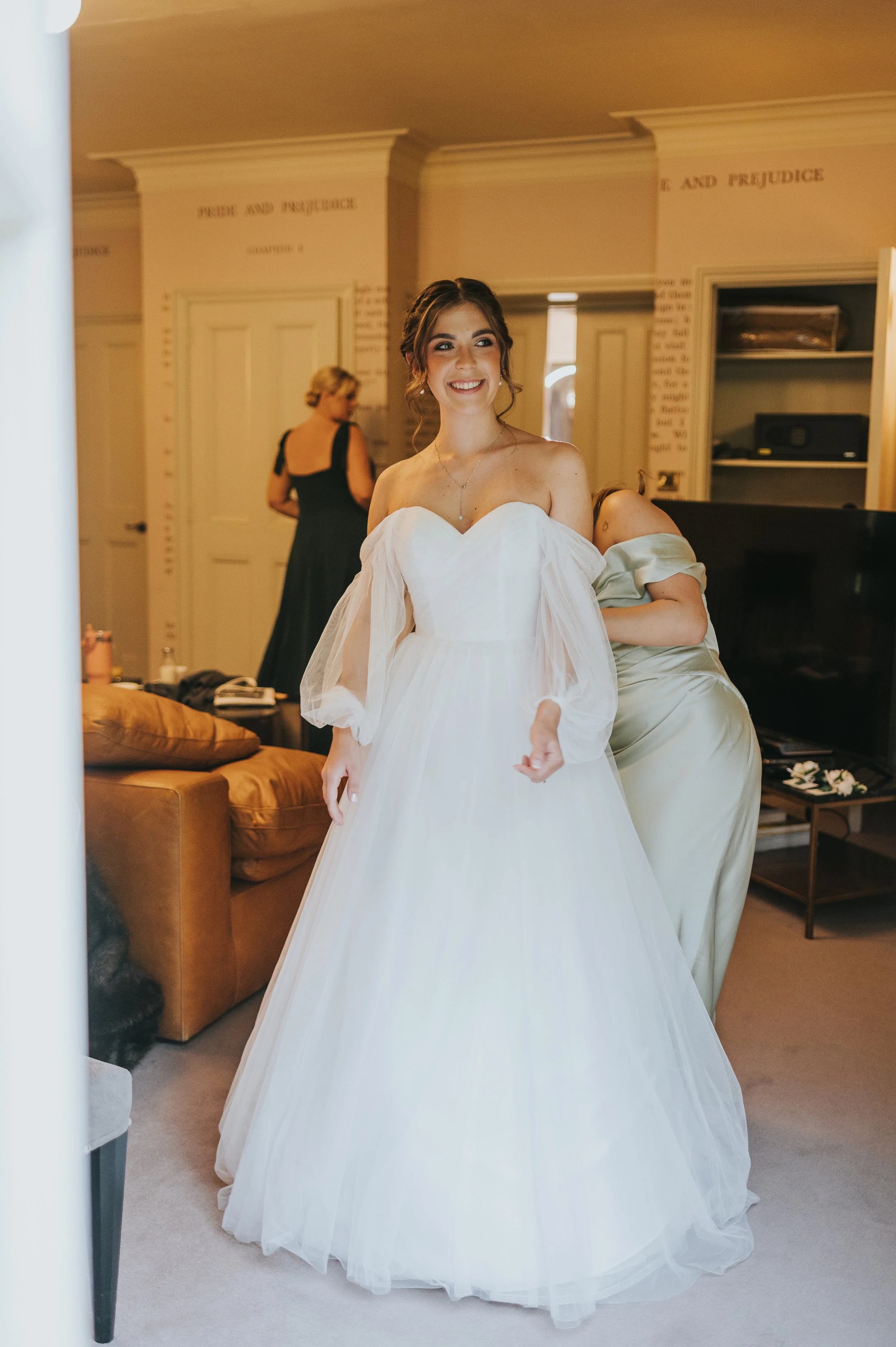 A young woman in a white wedding dress smiling in a room with other women preparing for a wedding. Rebecca Louise Photography Essex Wedding photographer