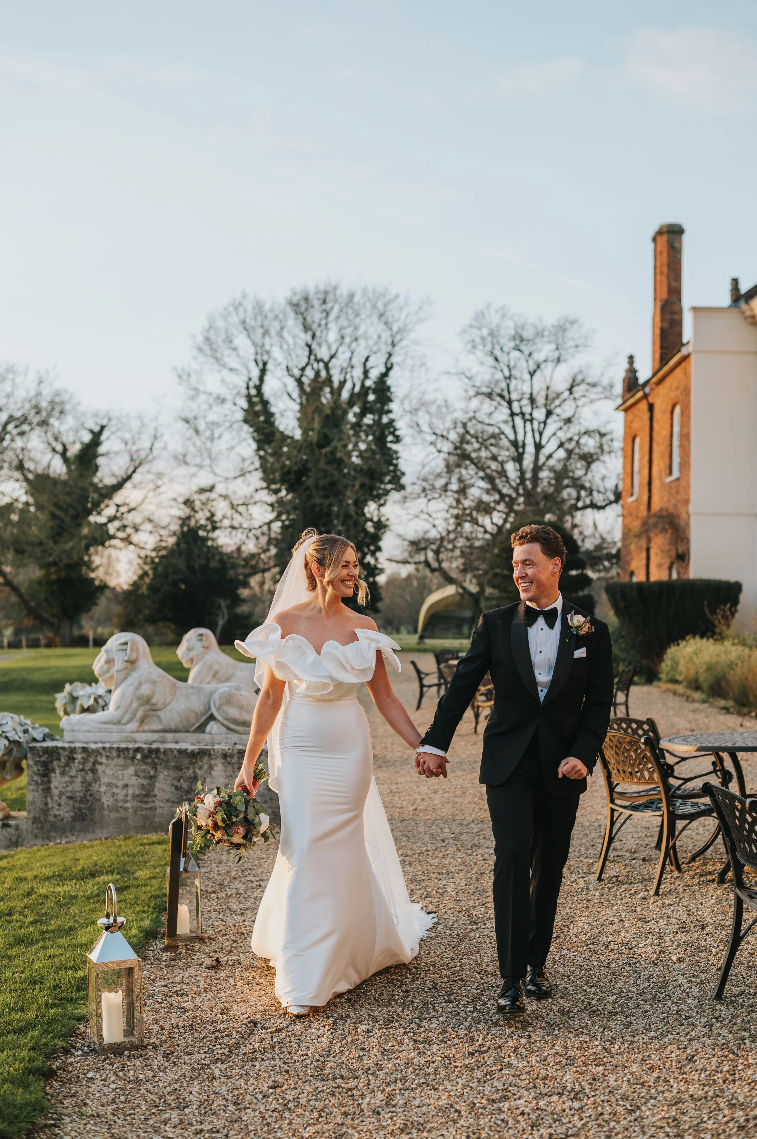 Happy bride and groom walking hand in hand outdoors during their wedding, smiling and looking at each other, surrounded by decorative lanterns and a scenic garden setting.