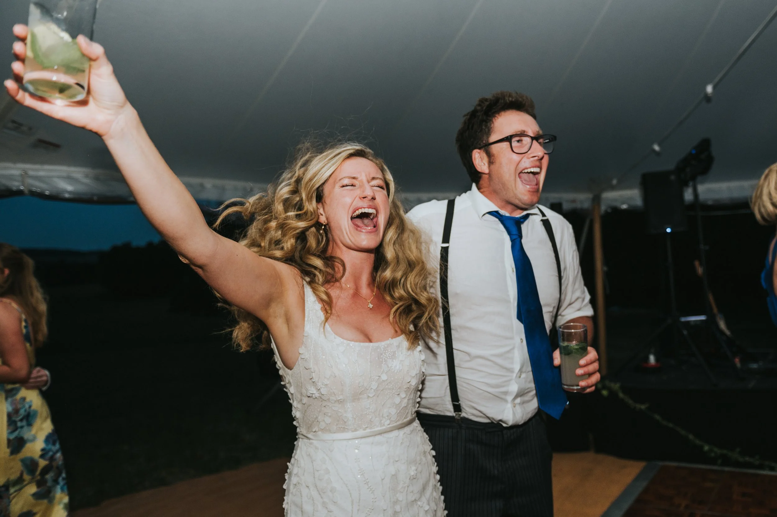 A joyous woman in a white wedding dress dancing with a man in a white shirt, blue tie, and suspenders at an outdoor celebration under a tent. She has her arm raised with a drink, both are smiling and singing.
