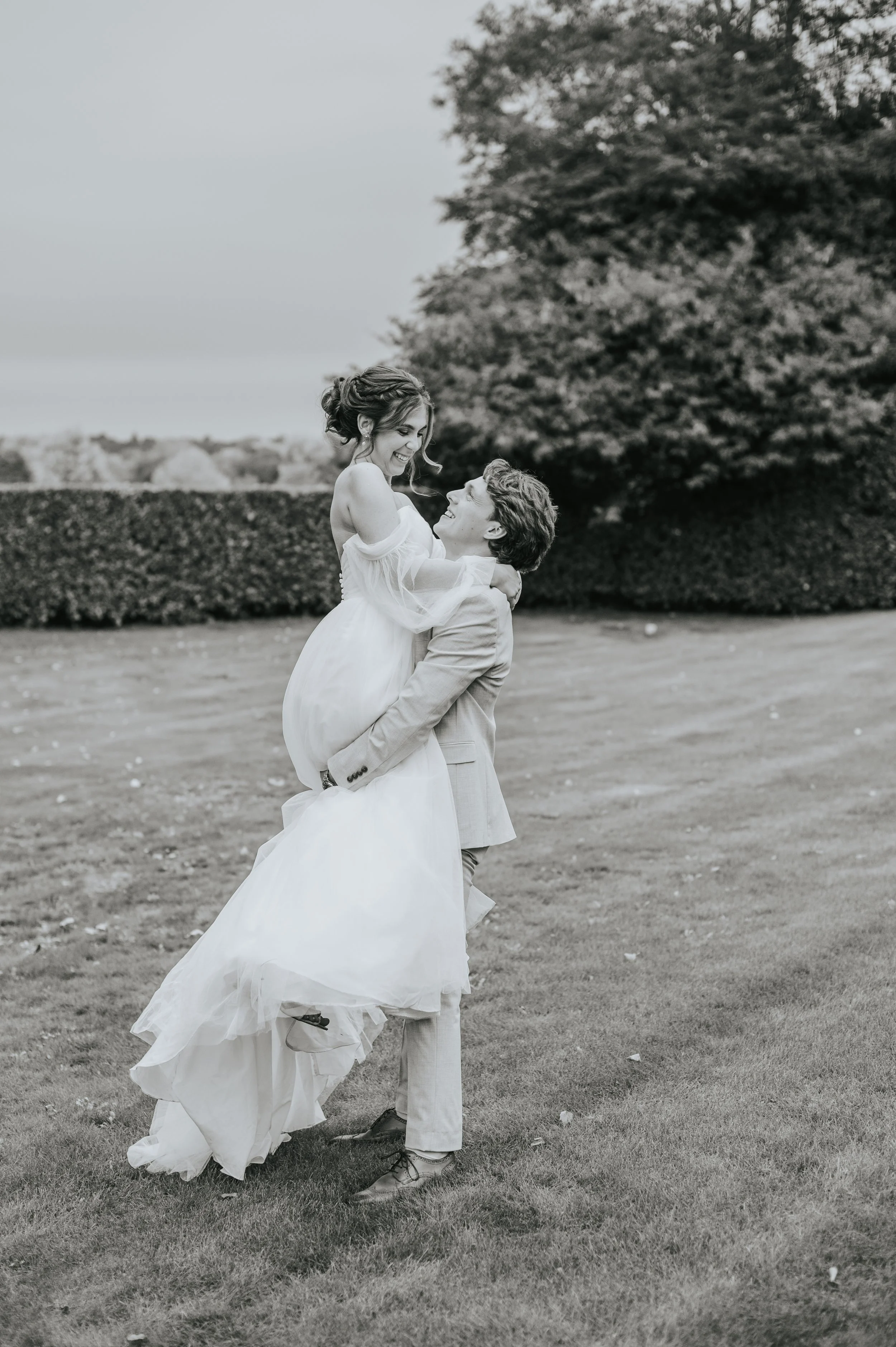 A happy couple on their wedding day outdoors, with the groom lifting the bride, both smiling and looking at each other, in a grassy area with trees and hedges in the background.