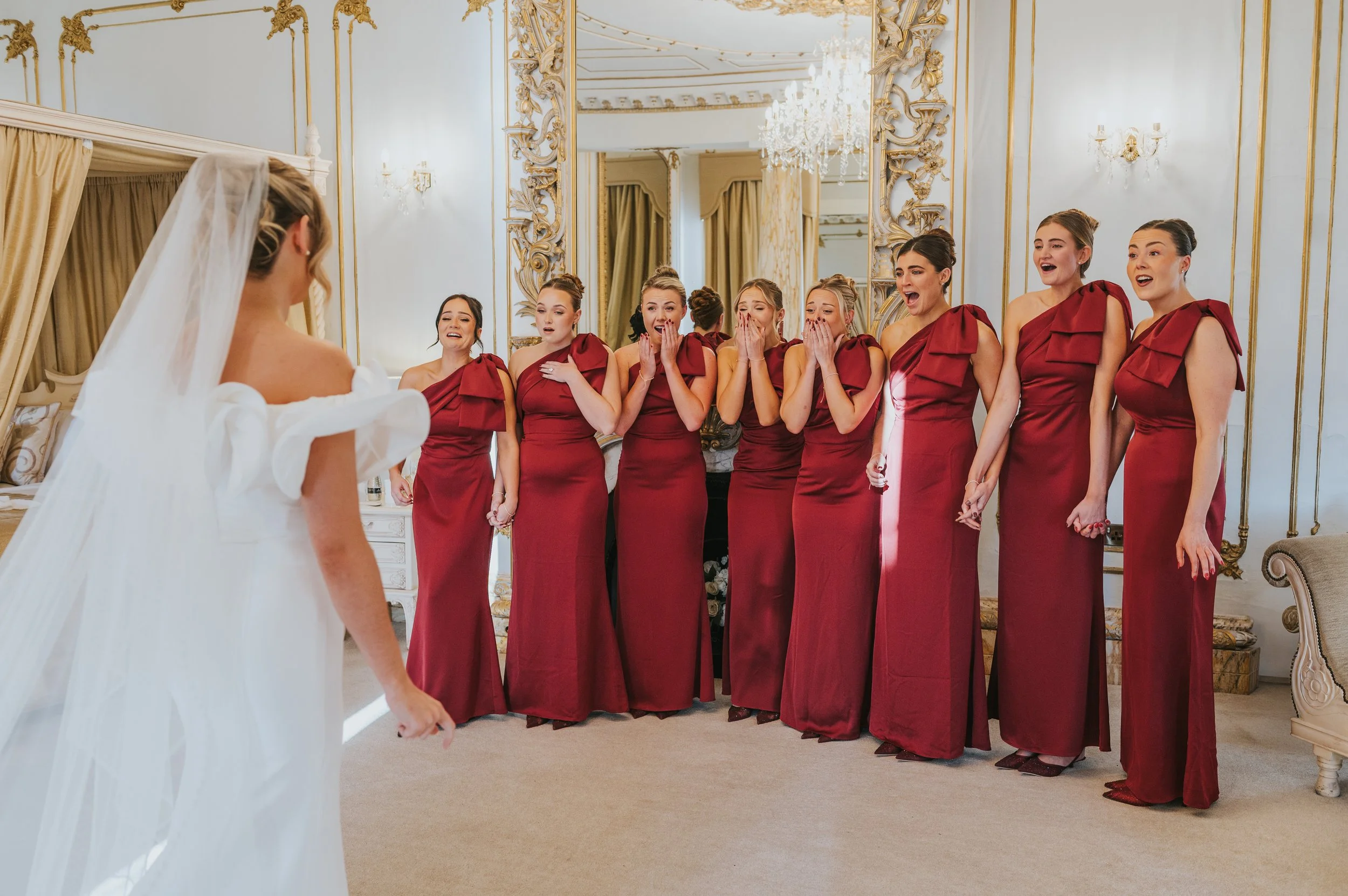 A bride in a white wedding dress and veil looking at a group of nine bridesmaids in matching red dresses, with emotional reactions, inside an ornate room with gold accents and a chandelier.