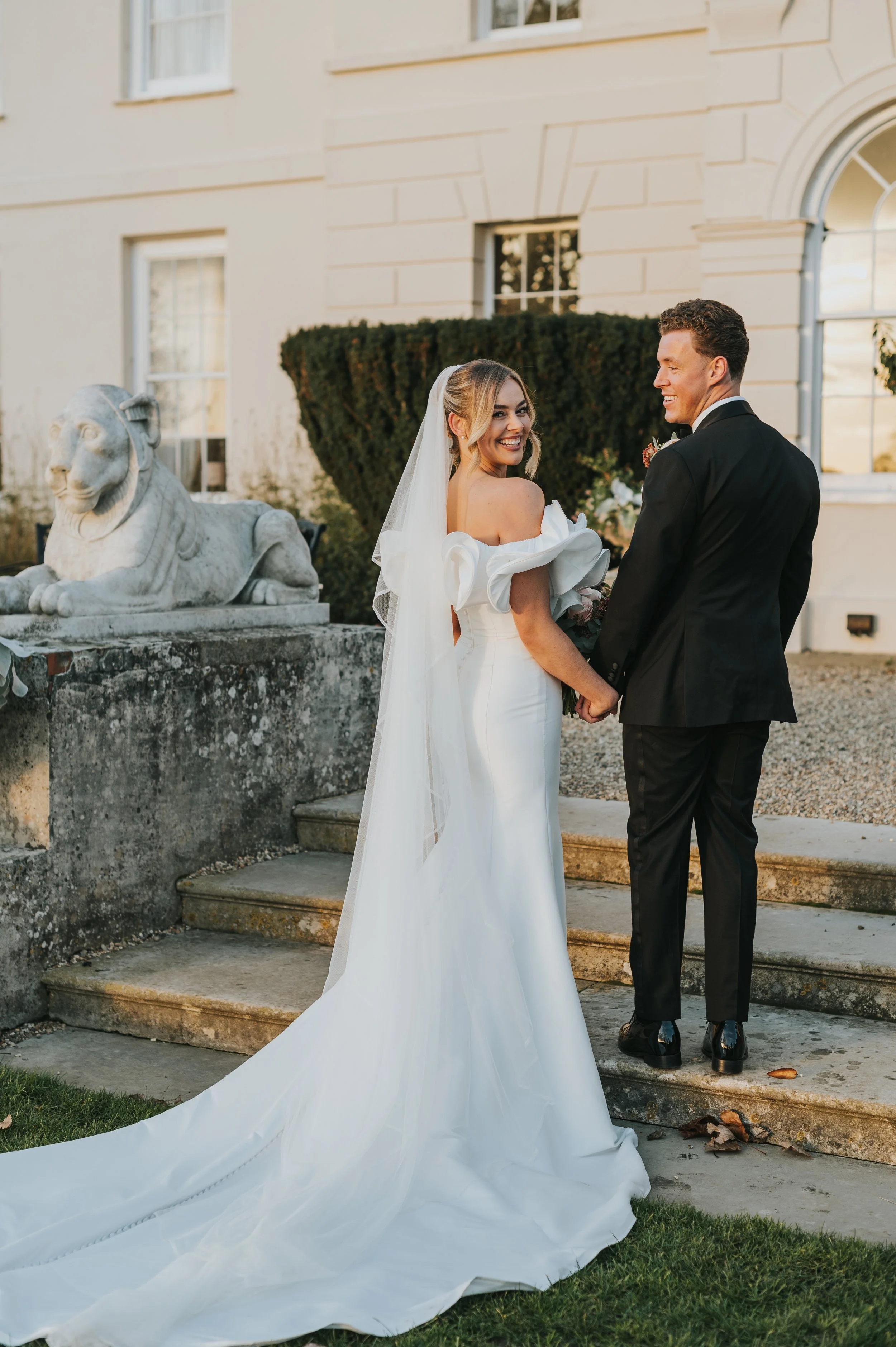 A bride and groom holding hands outside a building with stone stairs, a lion statue, and bushes, during sunset. The bride is wearing a white wedding gown with puffy off-shoulder sleeves and a long train, and the groom is in a black suit with a white 