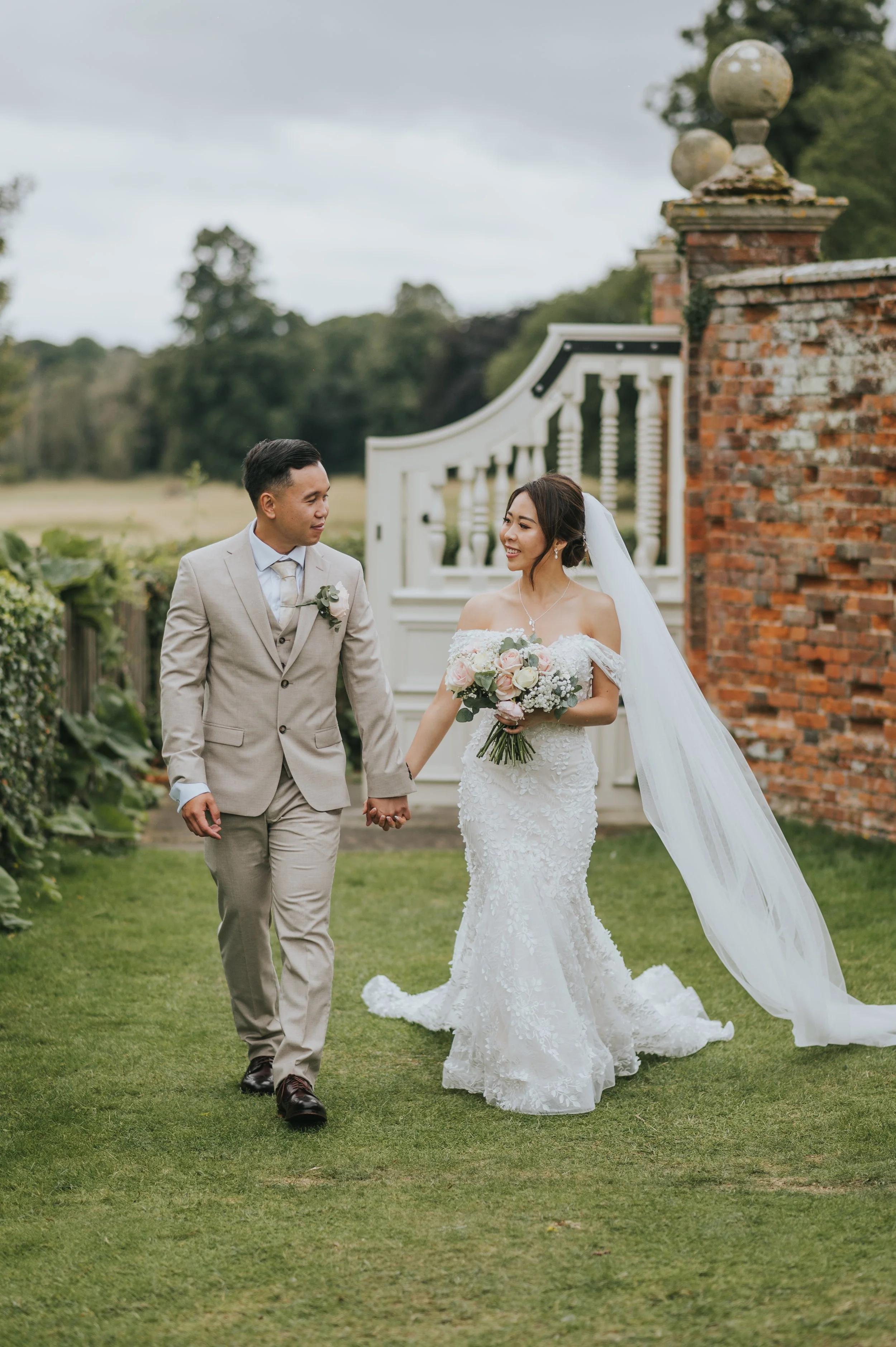 A bride and groom hold hands and walk on a grassy area outdoors, with the bride wearing a white lace wedding dress and veil, and the groom in a light beige suit, during a wedding photoshoot.